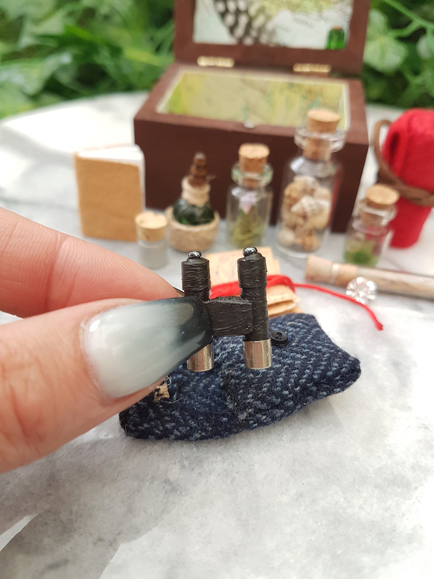 Hand holds a pair of tiny black binoculars along with a tiny tweed pouch. An open travellers chest sitsin the background glass vials, leather book, quill and inkwell, papers and red blanket roll. The inside of the chest is lined with map paper.