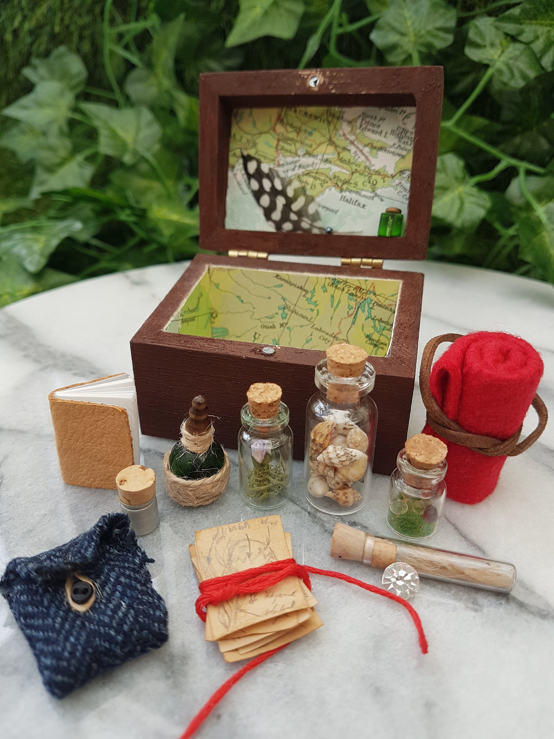 An open travellers chest is shown surrounded by filled glass vials, tweed pouch, leather book, papers and blanket roll. The inside of the chest is lined with coloured map paper