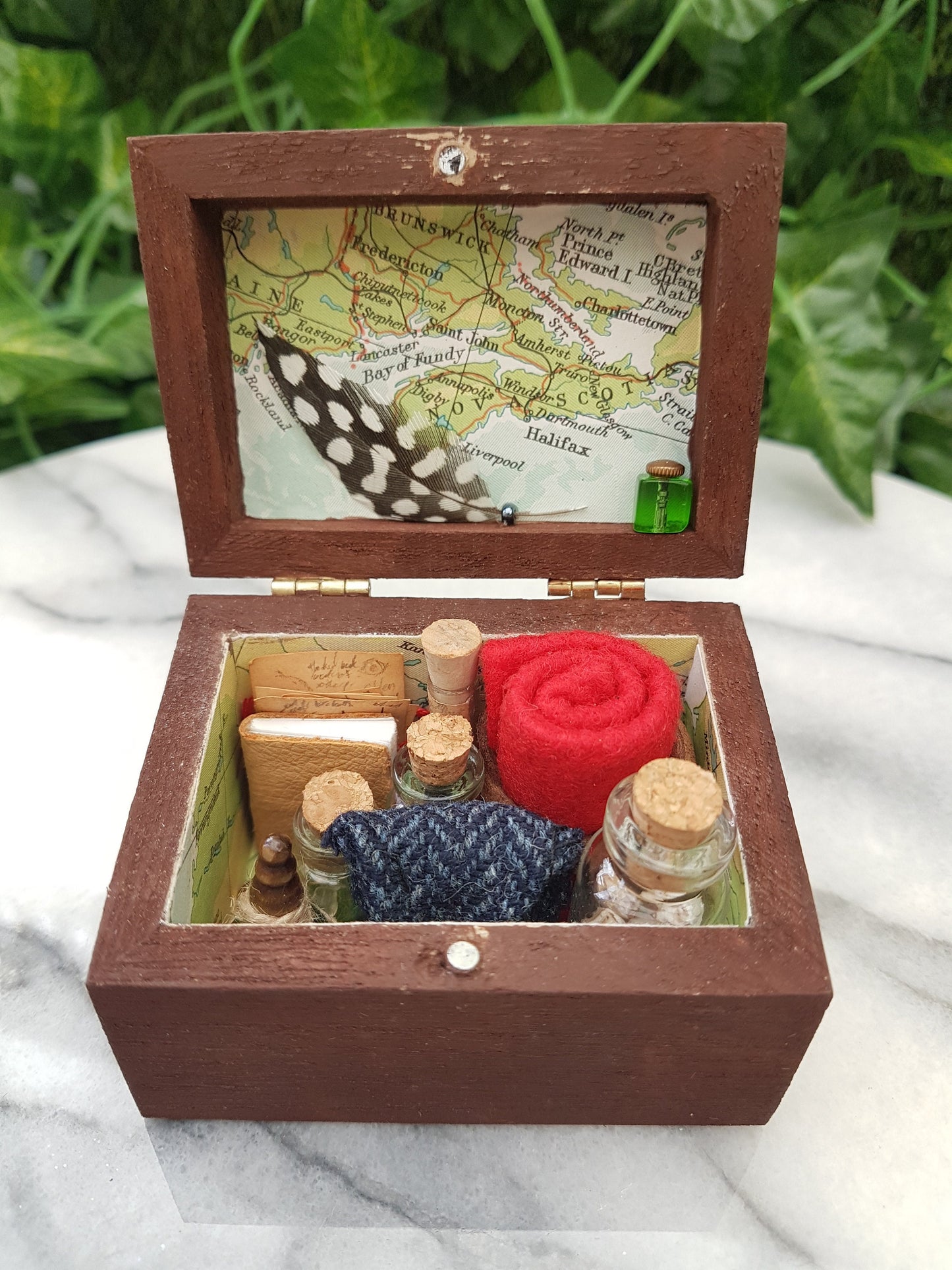 An open travellers chest is shown filled neatly with glass vials, tweed pouch, leather book, quill and inkwell, papers and red blanket roll. The inside of the chest is lined with coloured map paper. Shown on a marble slab