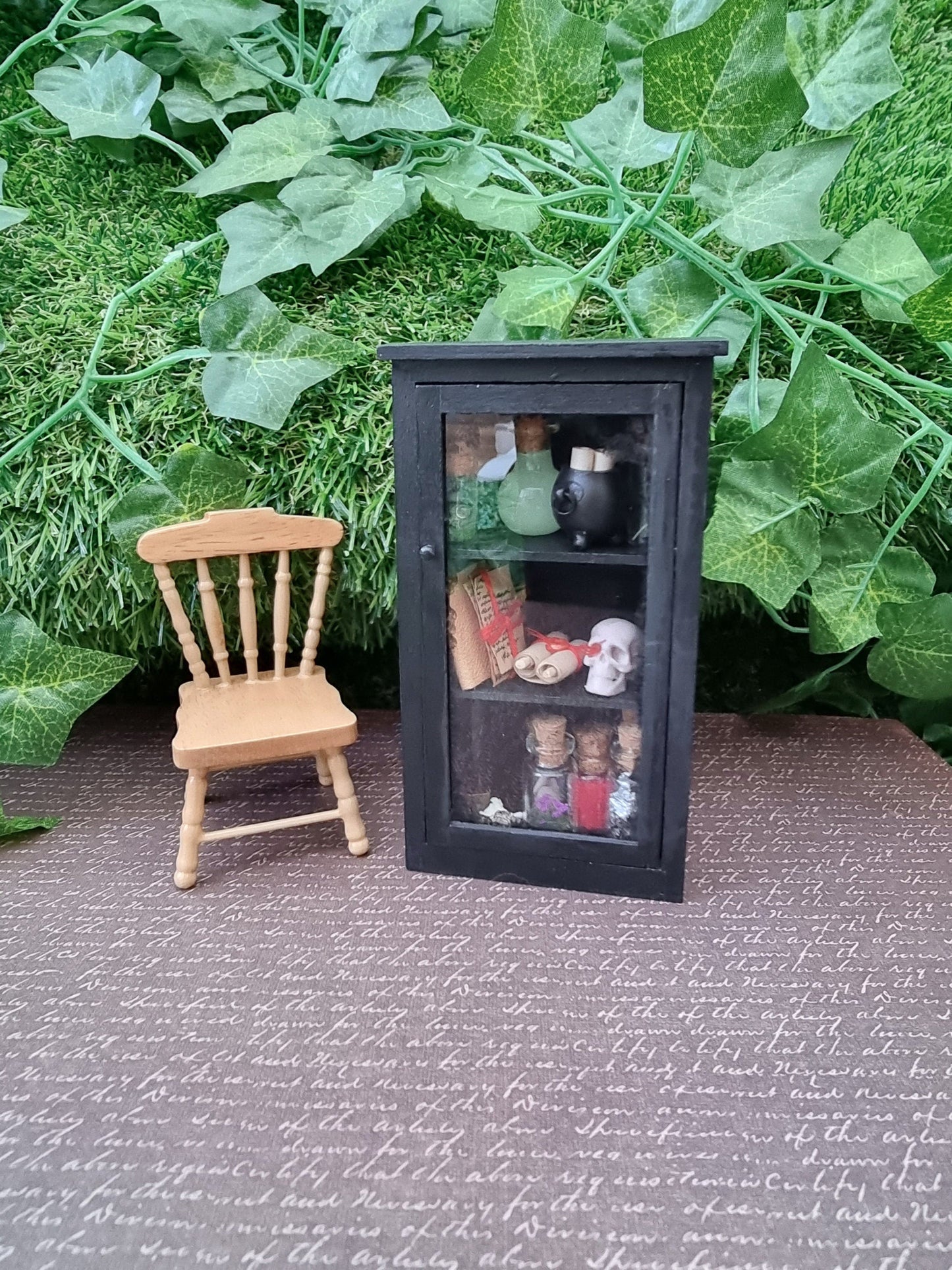 A miniature 1:12 scale glass-fronted black apothecary cabinet stands beside a minature chair against an ivy background. The cabinet has 3 shelves. The top shelf has apothecary bottles and a cauldron. Middle shelf has a skull and scroll. Bottom shelf has bottles