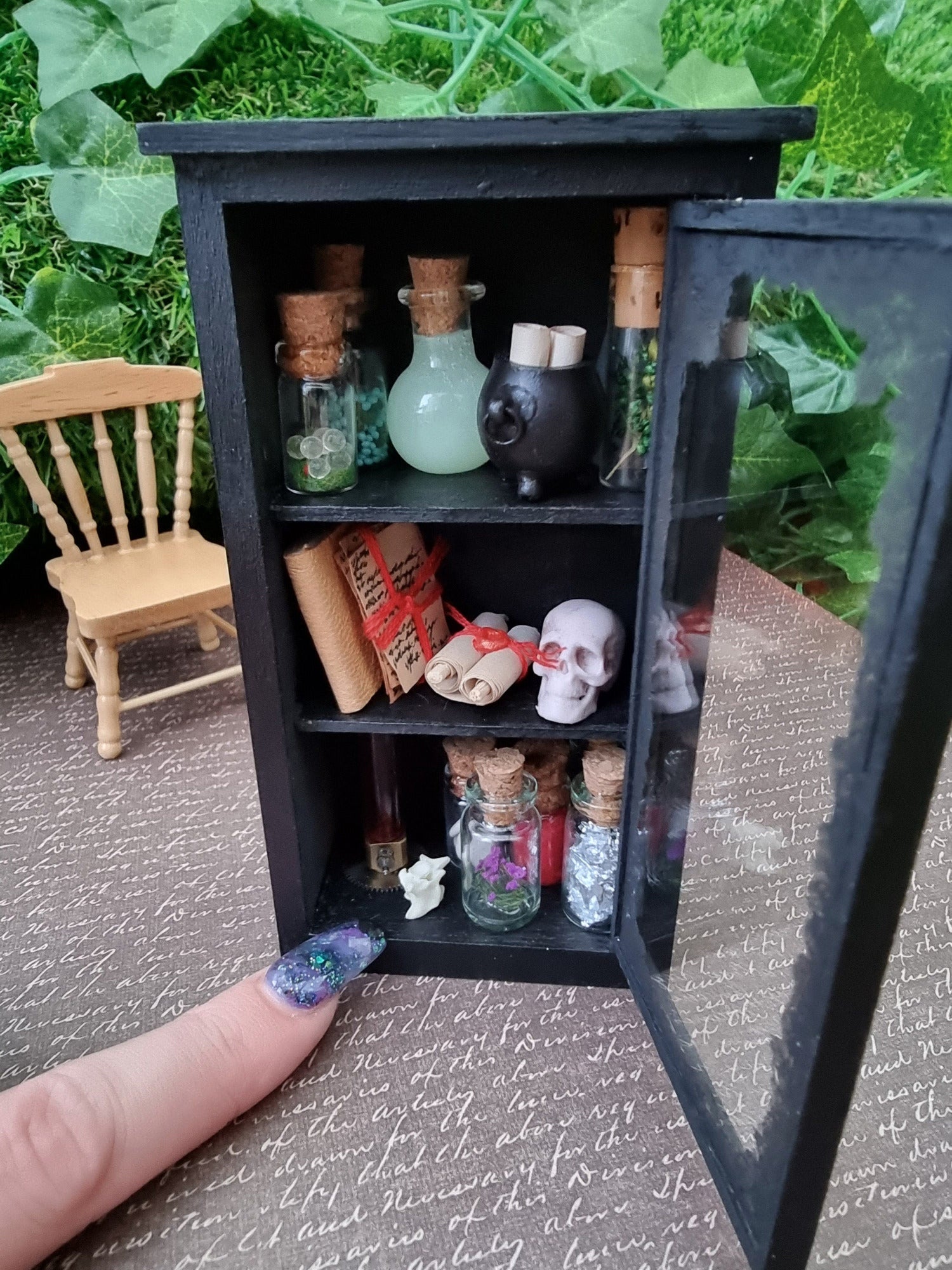 A miniature 1:12 scale glass-fronted black apothecary cabinet stands against an ivy background. The cabinet has 3 shelves. The top shelf has apothecary bottles and cauldron. Middle shelf has a skull and scroll. Bottom shelf has bottles. Finger points for scale