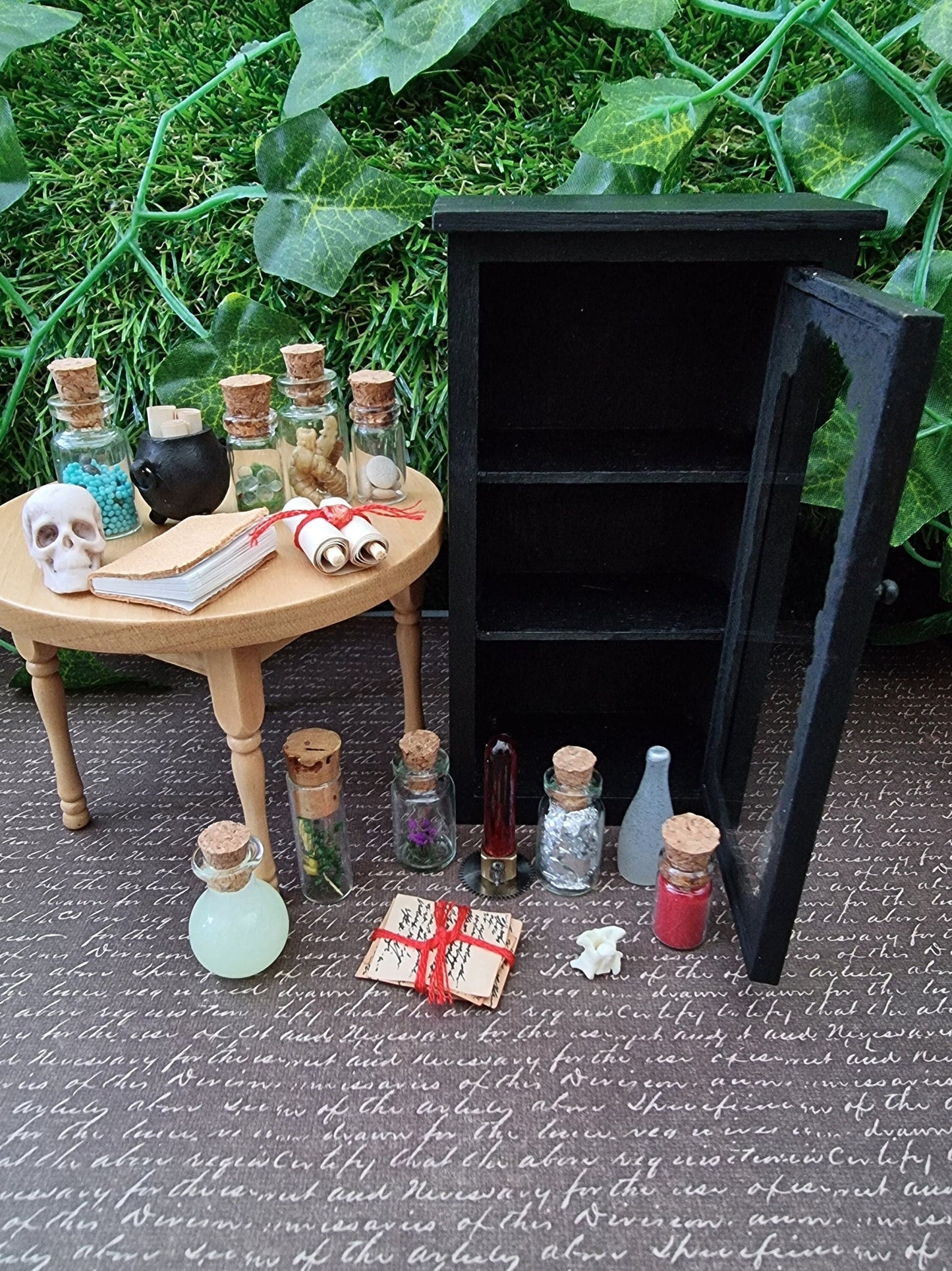 Open miniature 1:12 scale glass-fronted black apothecary cabinet stands against an ivy background. The cabinet has 3 shelves and is open and empty with contents sat beside, including apothecary bottles, a cauldron, book, skull and scroll.