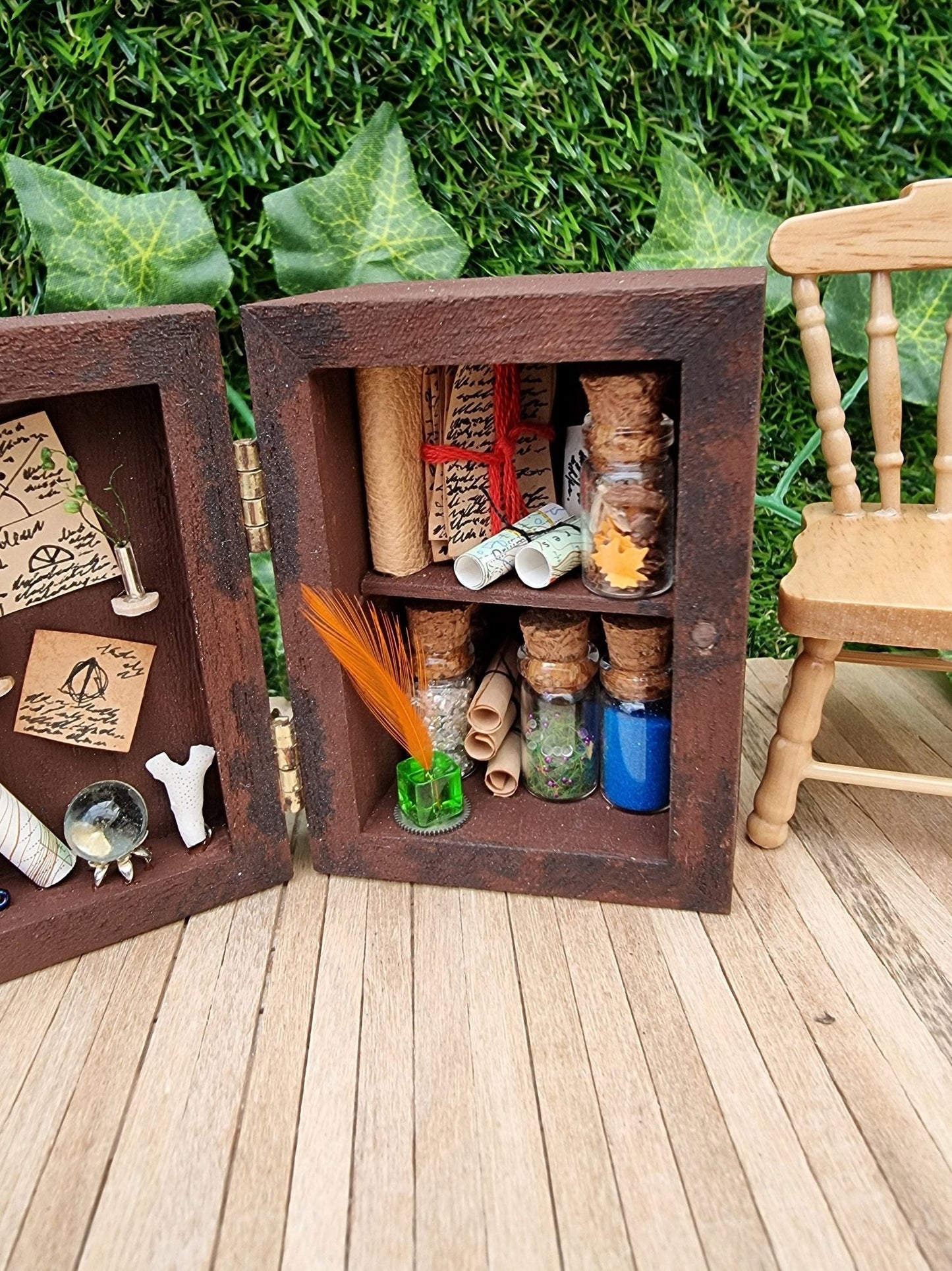 A 1:12 scale miniature brown apothecary cabinet shown open on a wood floor containing glass vials, parchment scrolls, letters, notes, inkwell and quill. The left cabinet door has aged notes pinned up, shells, crystal ball, map, coral and slim vases