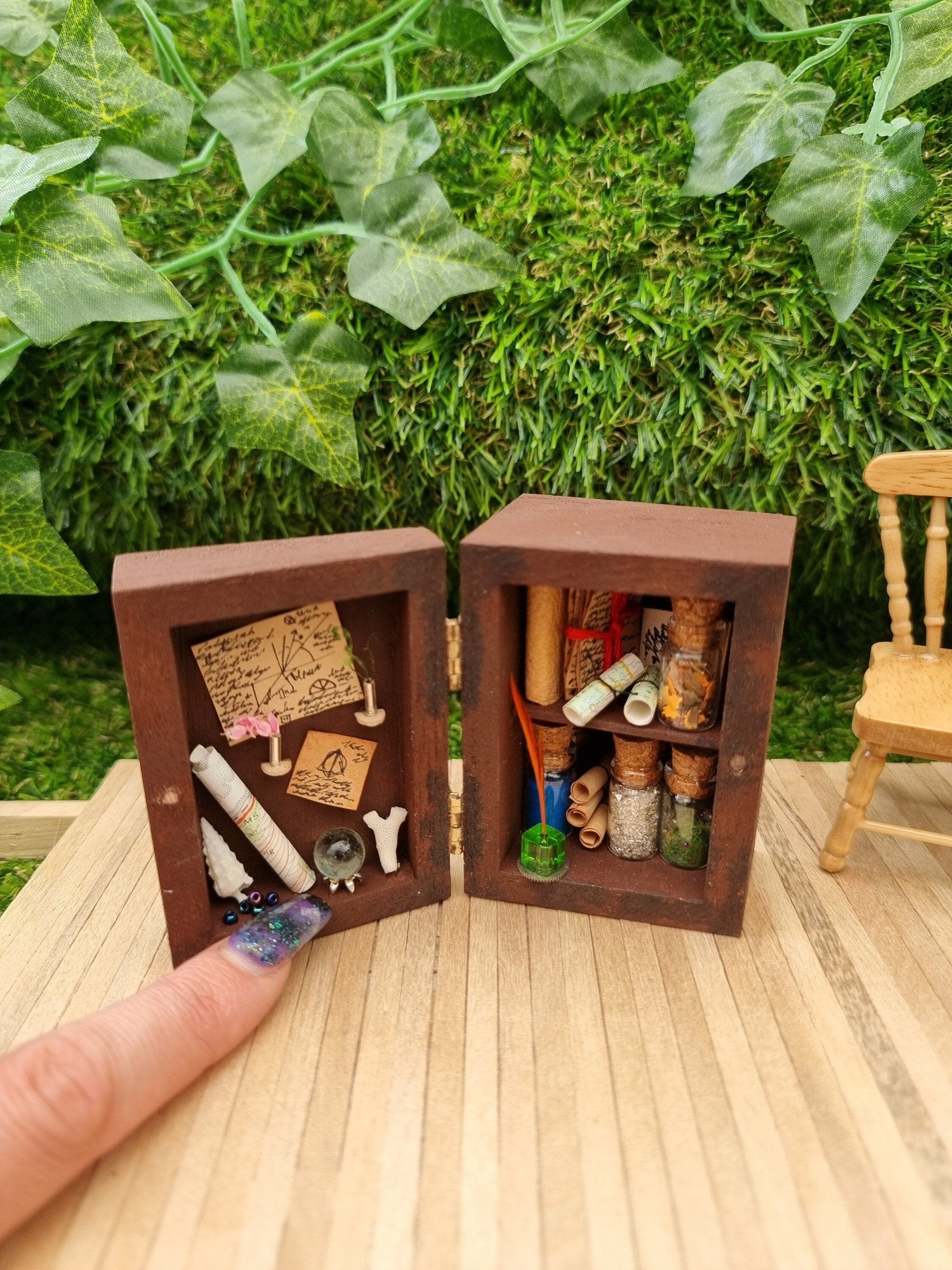 A 1:12 scale miniature brown apothecary cabinet shown open, containing glass vials, parchment scrolls, letters, notes, inkwell and quill. The left cabinet door has aged notes, shells, crystal ball, map, coral and vases. A finger points for scale