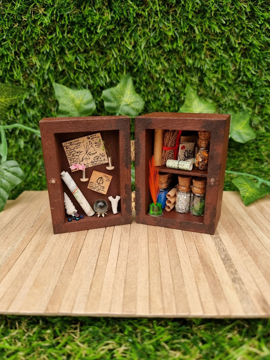 A 1:12 scale miniature brown apothecary cabinet is shown open on a marble slab with glass vials, parchment scrolls, miniature cauldron and leather bound book. The left cabinet door has aged notes pinned up with an inkwell and quill