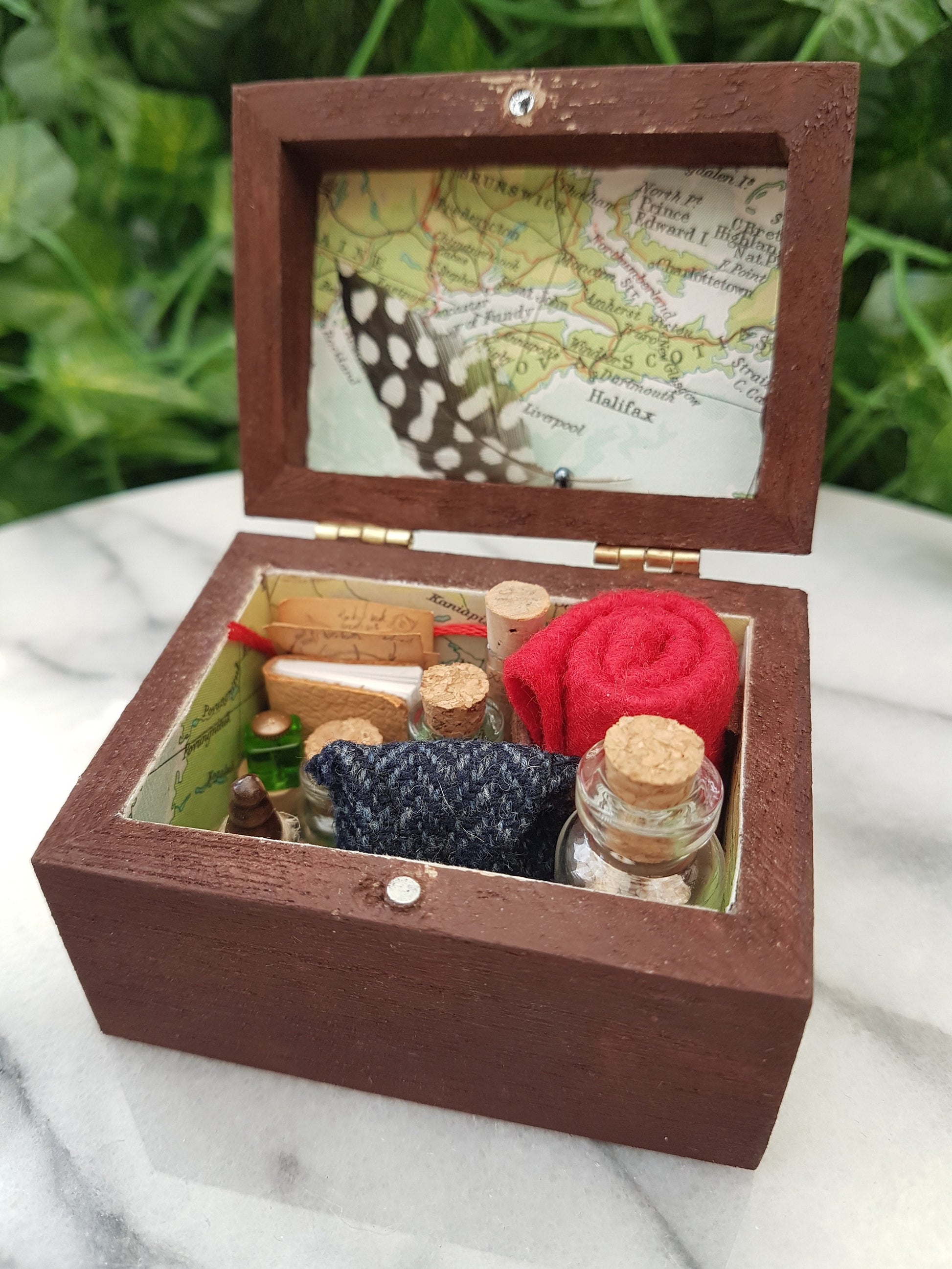 An open travellers chest is shown filled neatly with glass vials, tweed pouch, leather book, quill and inkwell, papers and red blanket roll. The inside of the chest is lined with coloured map paper. Shown on a marble slab