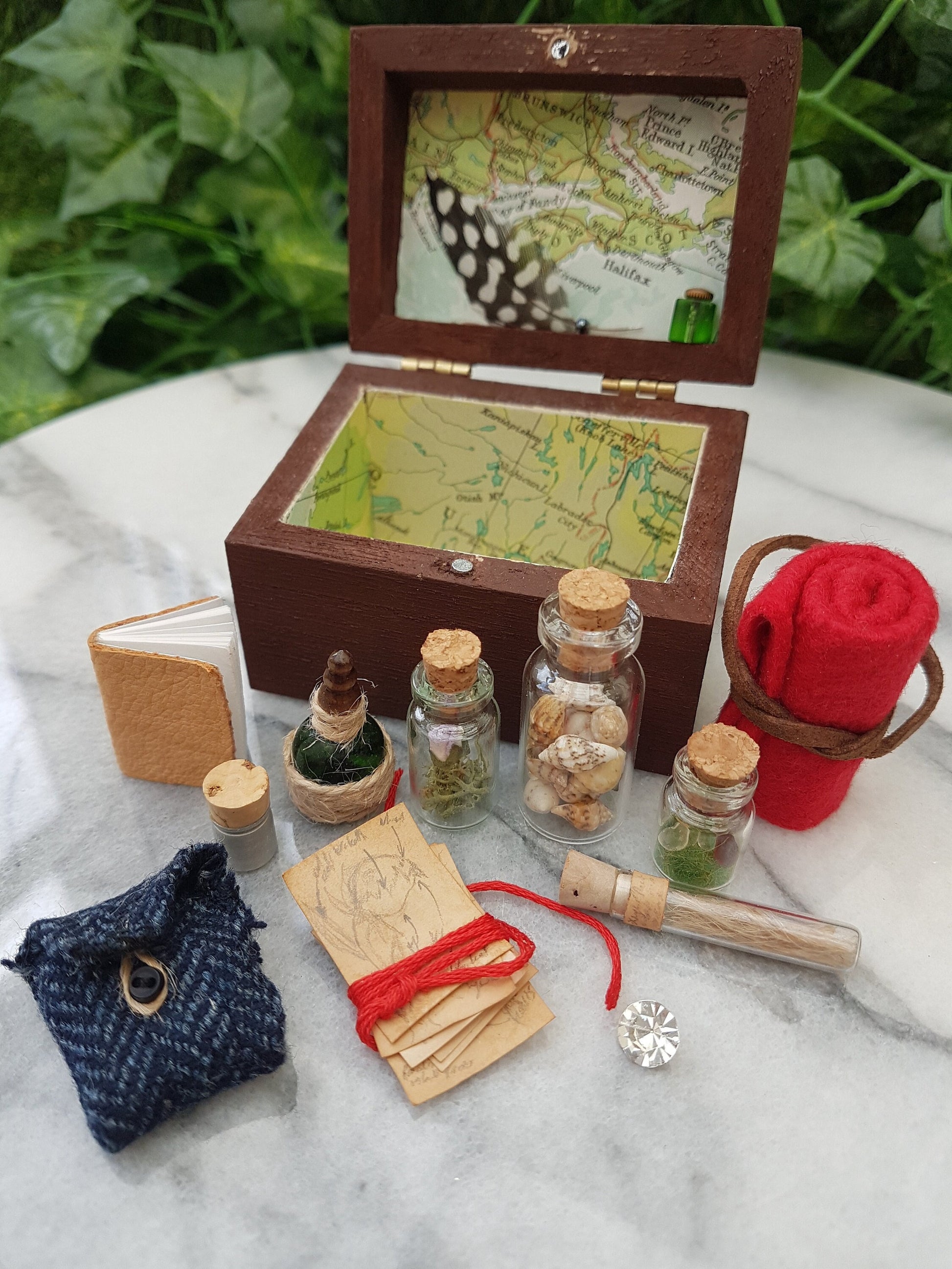 An open travellers chest is shown surrounded by filled glass vials, tweed pouch, leather book, papers and blanket roll. The inside of the chest is lined with coloured map paper