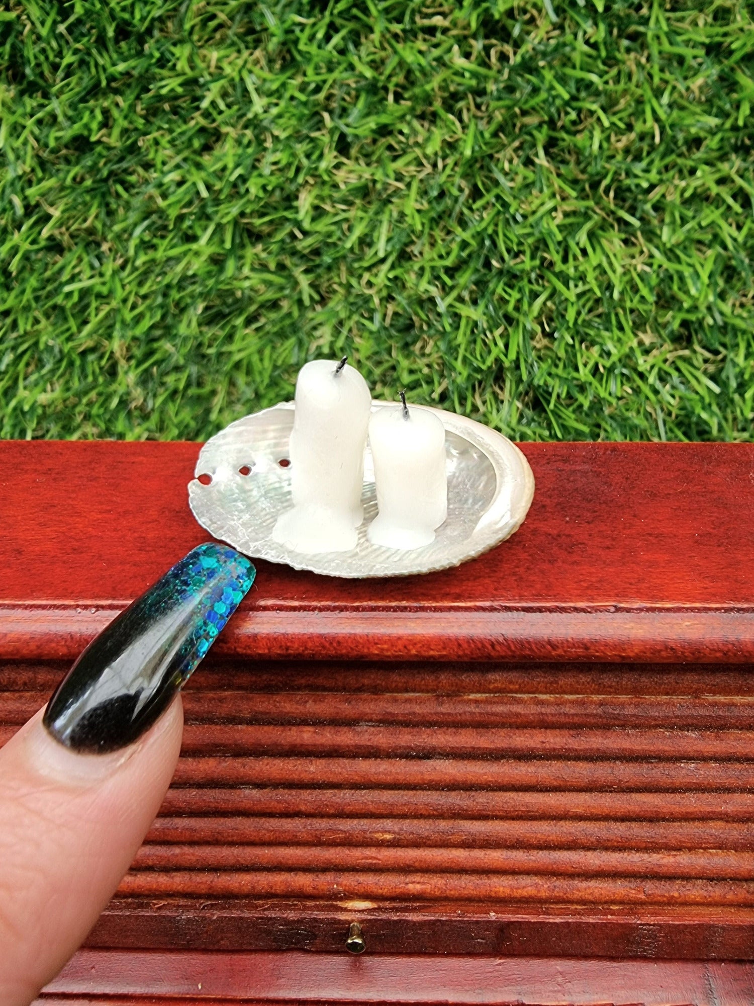 Two white 1:12 scale miniature candles in a small abalone shell dish, displayed on a miniature desk. A finger shows scale