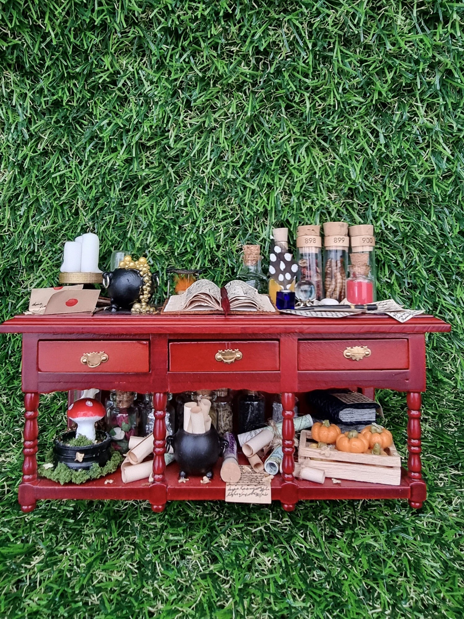 A 1:12 scale miniature brown wooden sideboard has 3 drawers and an open display area beneath. The bench top shows a bubbling cauldron, open spellbook, filled glass vials, parchments, wand, candles, burner, sand timer, mushroom garden and a crate of pumpkins