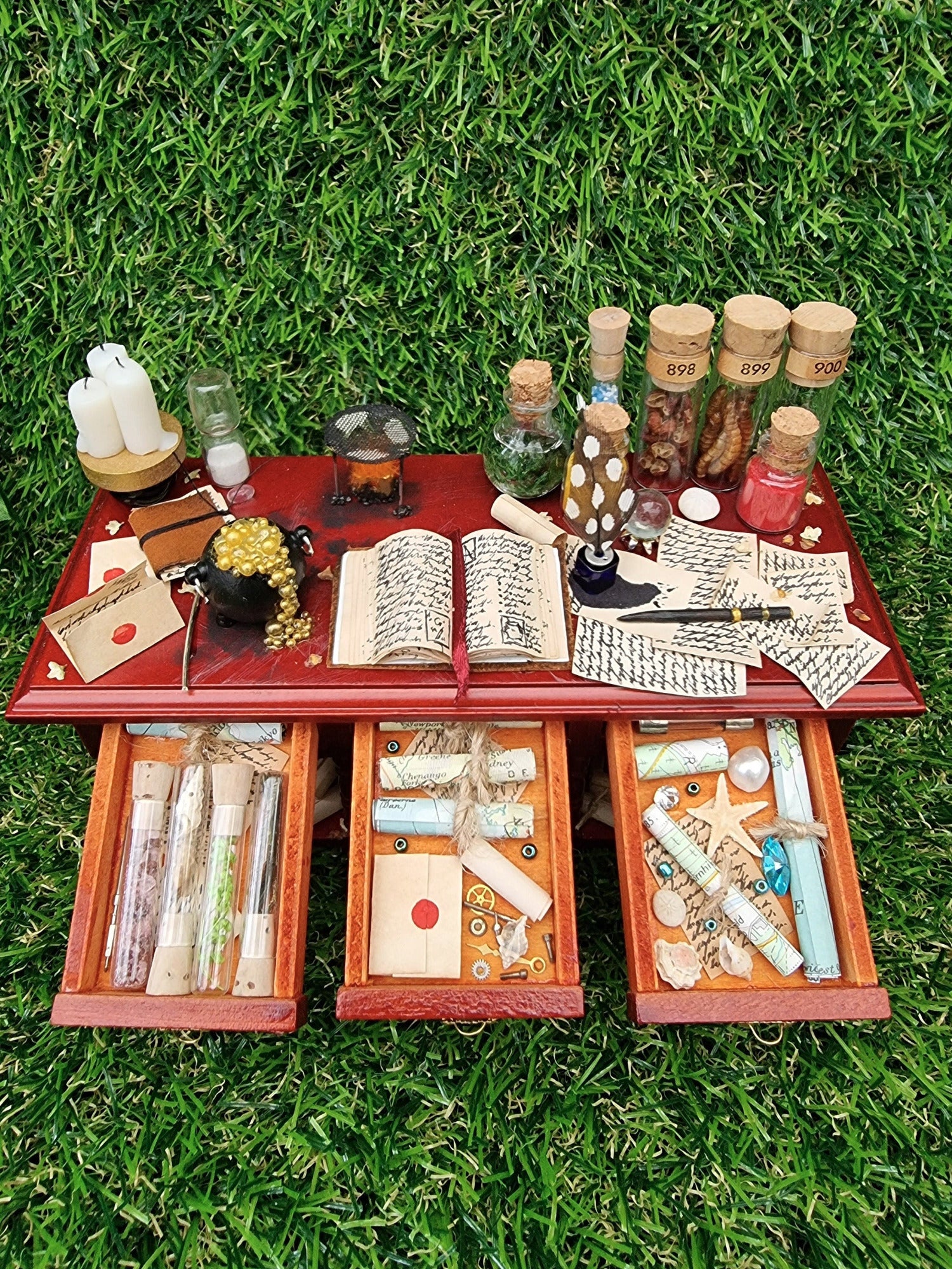 A miniature brown wooden sideboard has 3 open drawers. The bench top shows a bubbling cauldron, open spellbook, filled glass vials, parchments, wand, candles, burner, sand timer, and the drawers contain glass vials, letters, maps rolls and starfish