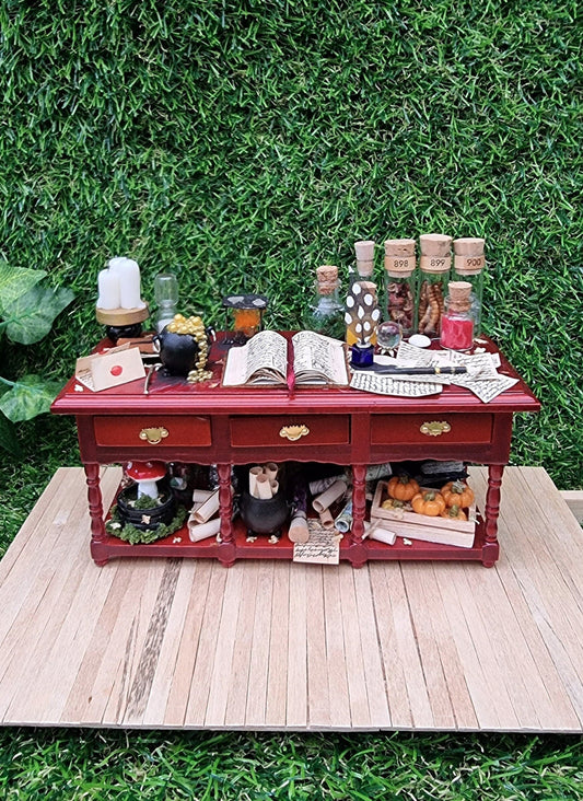 A 1:12 scale miniature brown wooden sideboard has 3 drawers and an open display area beneath. The bench top shows a bubbling cauldron, open spellbook, filled glass vials, parchments, wand, candles, burner, sand timer, mushroom garden and a crate of pumpkins