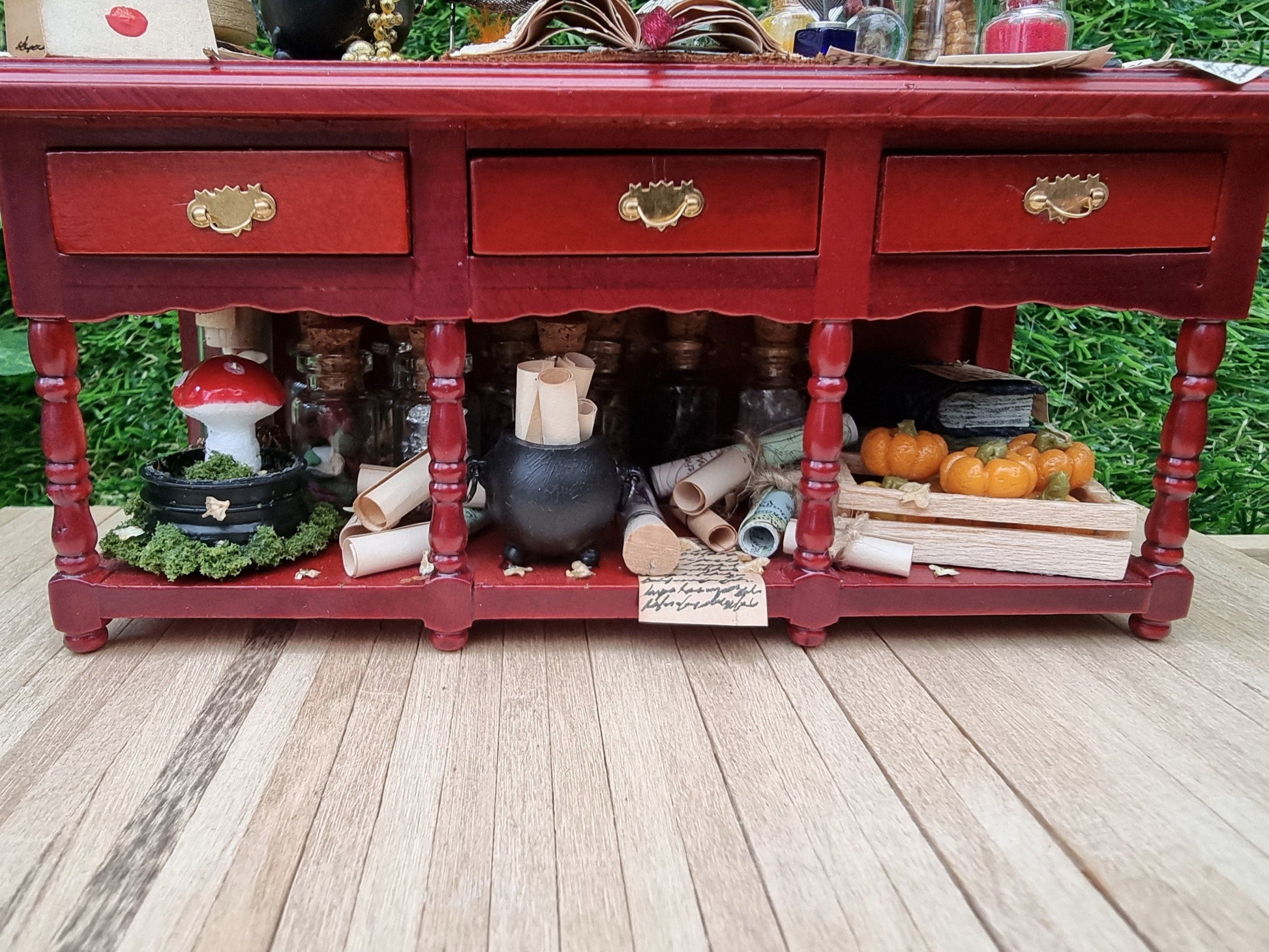 A 1:12 scale miniature brown wooden sideboard with 3 drawers and an open display area beneath. The display area shows a black cauldron with parchment rolls inside, filled glass vials, mushroom garden, maps rolls and a crate of pumpkins