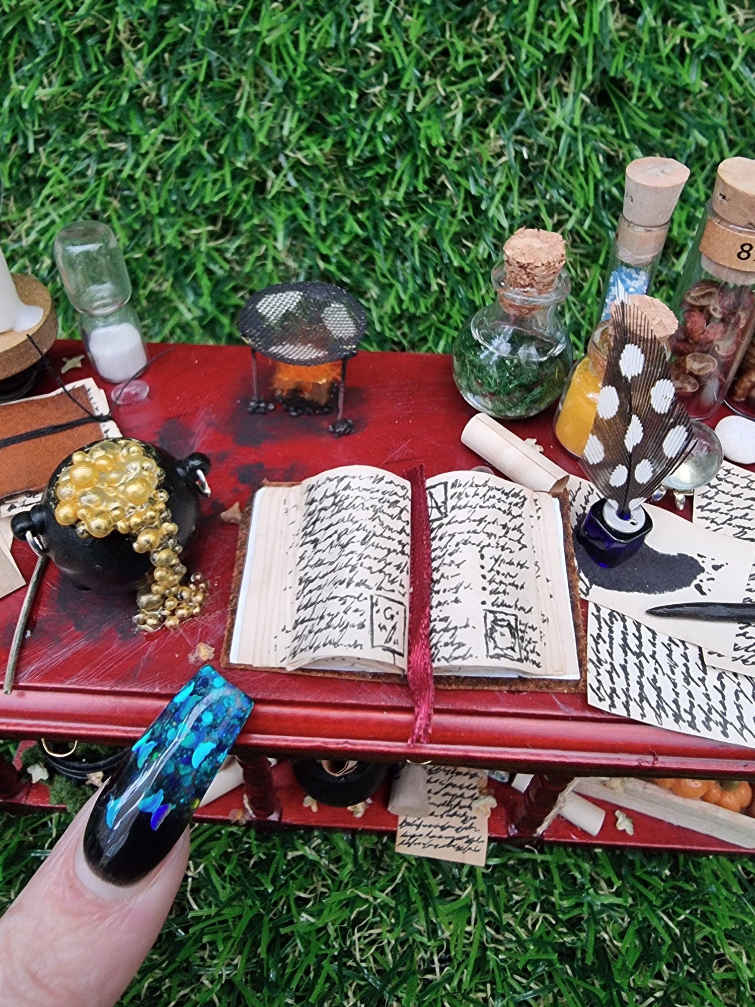 A finger points to a 1:12 miniature bench top at a tiny open spellbook, surrounded by bubbling black cauldron, burner, journal, sand timer, inkwell and quill, filled glass vials and parchments with an ink spill