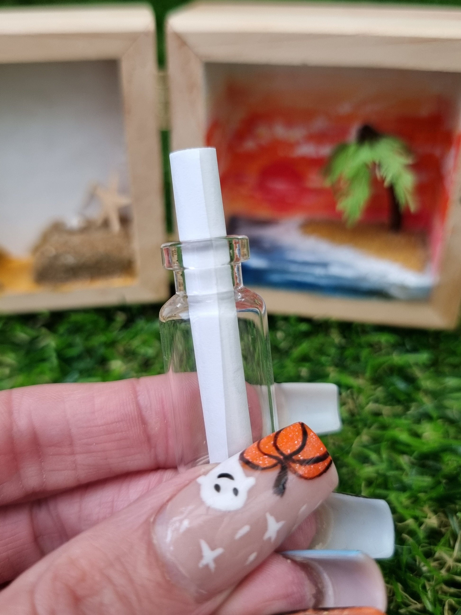 A hand holds a miniature glass bottle with a rolled up scroll ofwhite paper inside. A miniature diorama of a desert island sits in the background