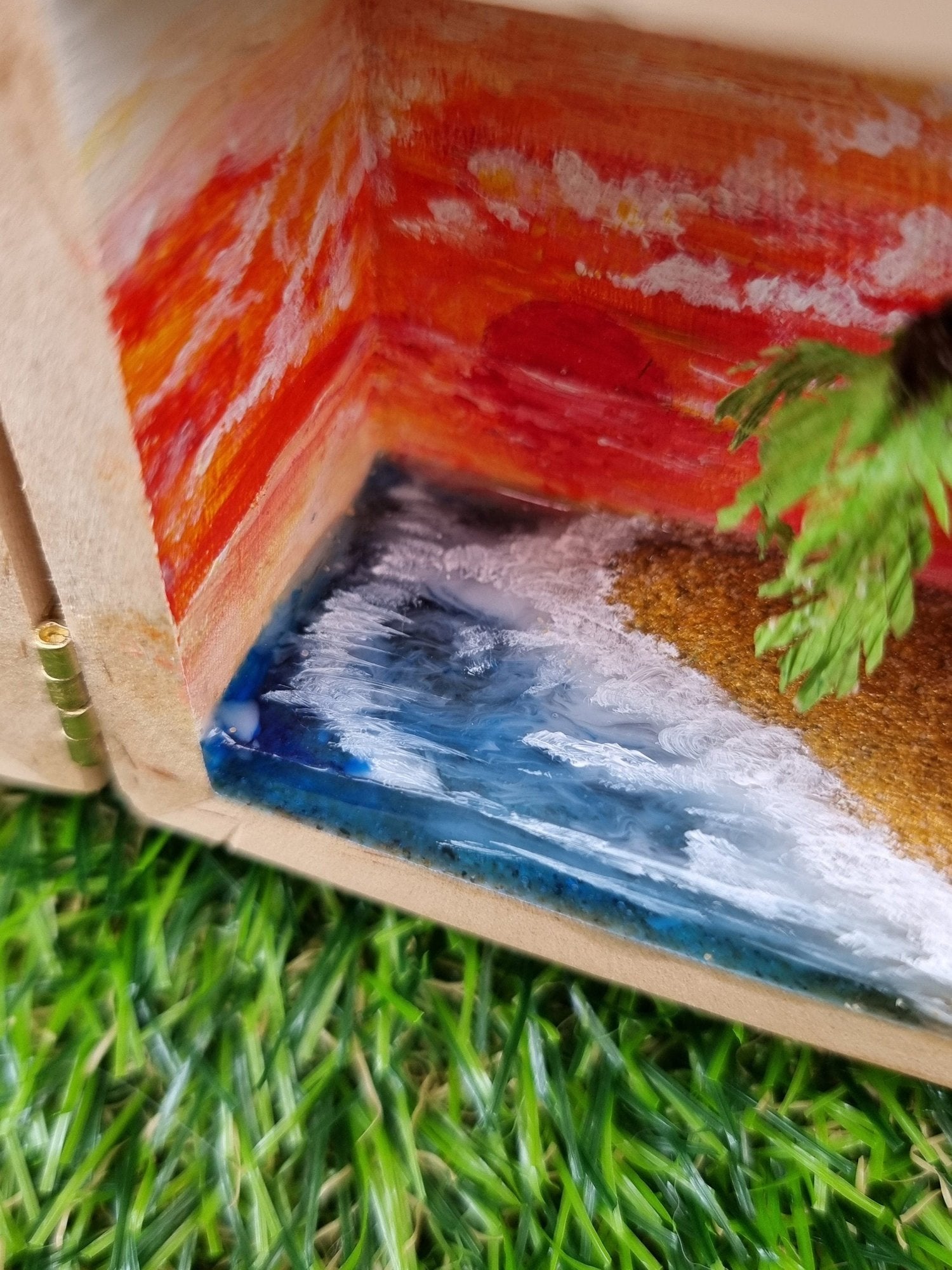 Detail of the right side of a box diorama, showing a miniature desert with a palm tree island surrounded by a deep blue resin sea  with white waves and foam lapping on the beach beneath an orange sunset