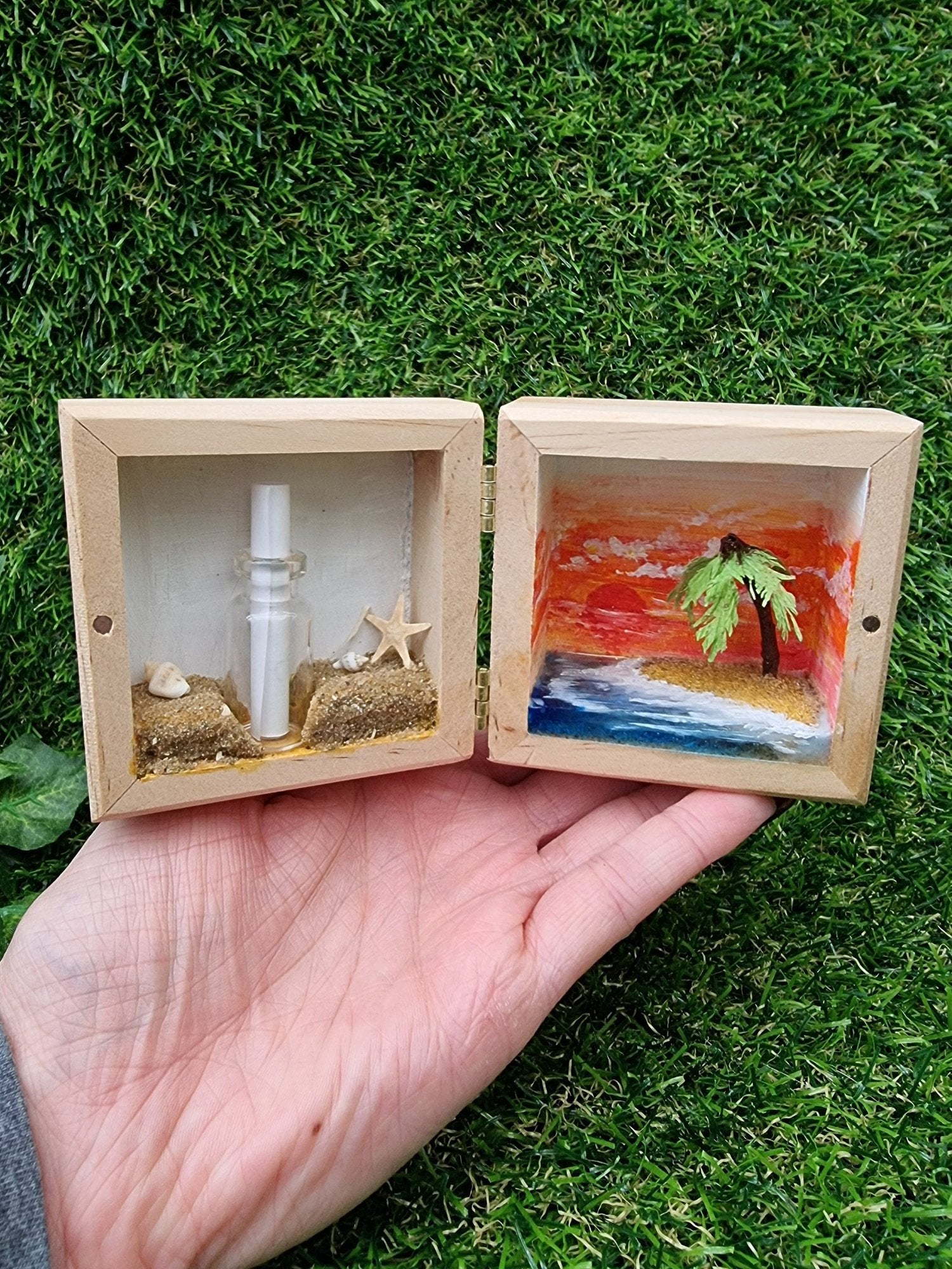 A hand holds a small open wooden box open like a book. The left side has a message in a bottle on a beach. The right side has a miniature desert with a palm tree island surrounded by blue sea and a deep orange sunset in the background