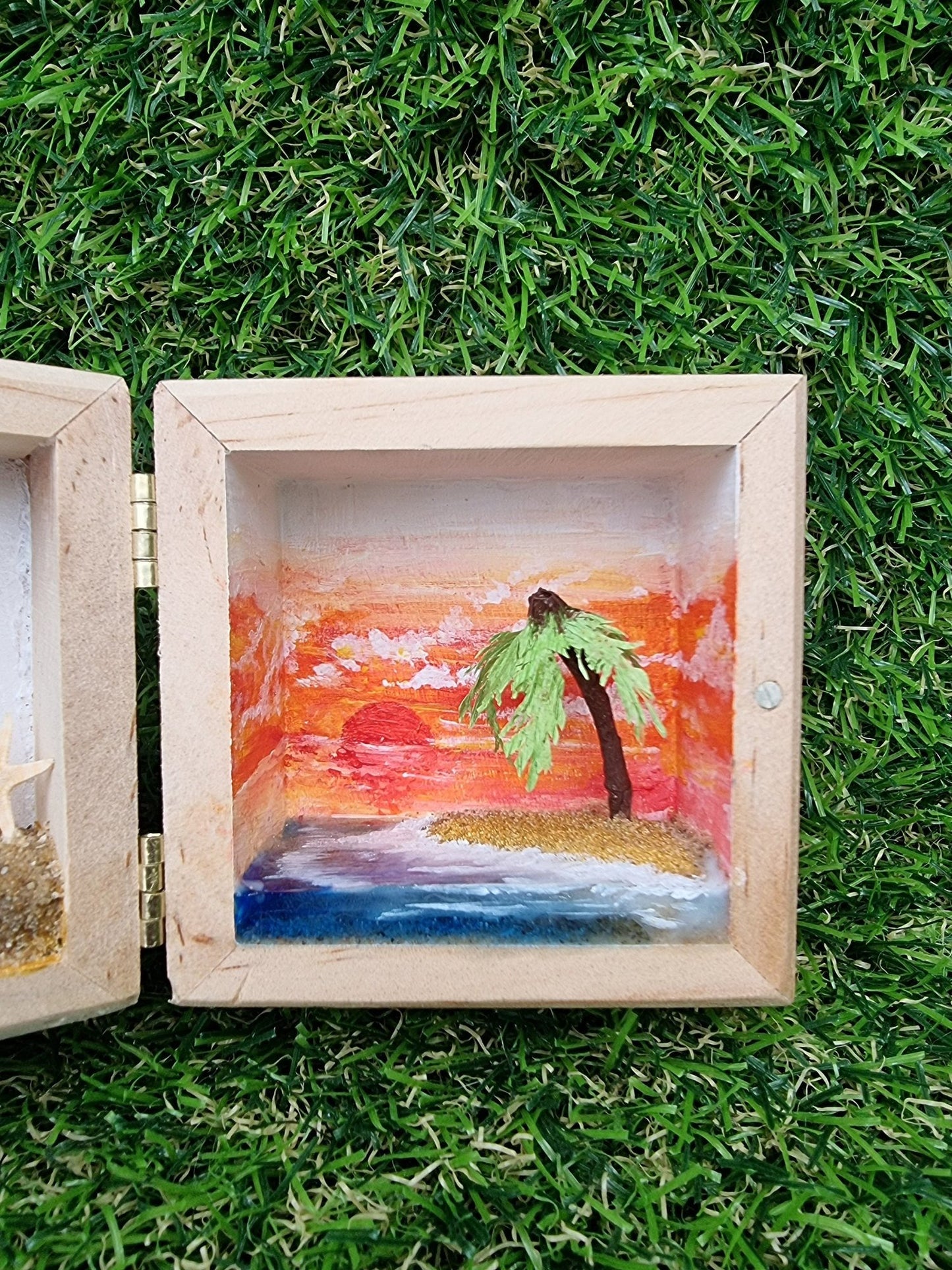 Detail of the right side of a box diorama, showing a miniature desert with a palm tree island surrounded by blue resin sea and a deep orange sunset in the background