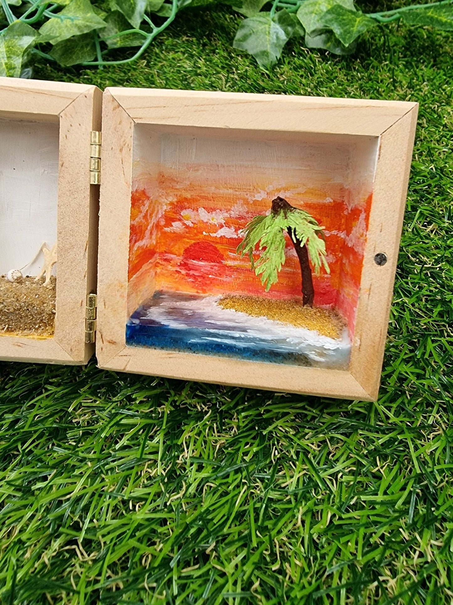 Detail of the right side of a box diorama, showing a miniature desert with a palm tree island surrounded by blue resin sea and a deep orange sunset in the background