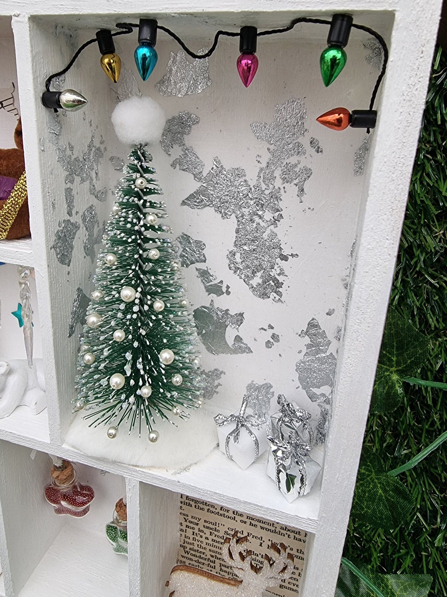 Detail of a small white shelving unit in the shape of a house. Each compartment has a different Christmas scene including christmas tree and presents with a silver leaf background and coloured christmas garland. A stag and baubles are seen beneath