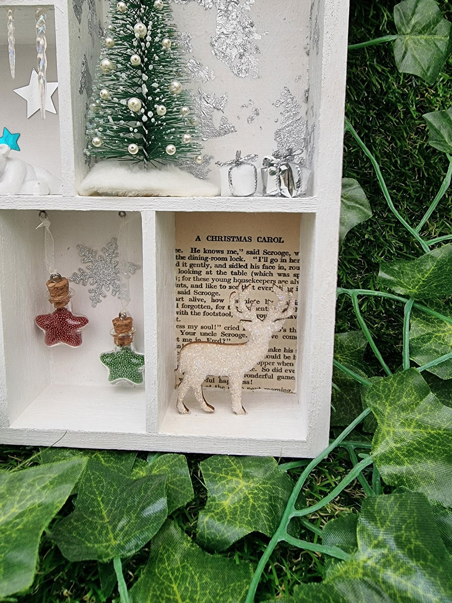 Detail of a small white shelving unit in the shape of a house. Each compartment has a different Christmas scene including hanging glass star baubles in green and red, presents and tree, and a stag with text background from a christmas carol