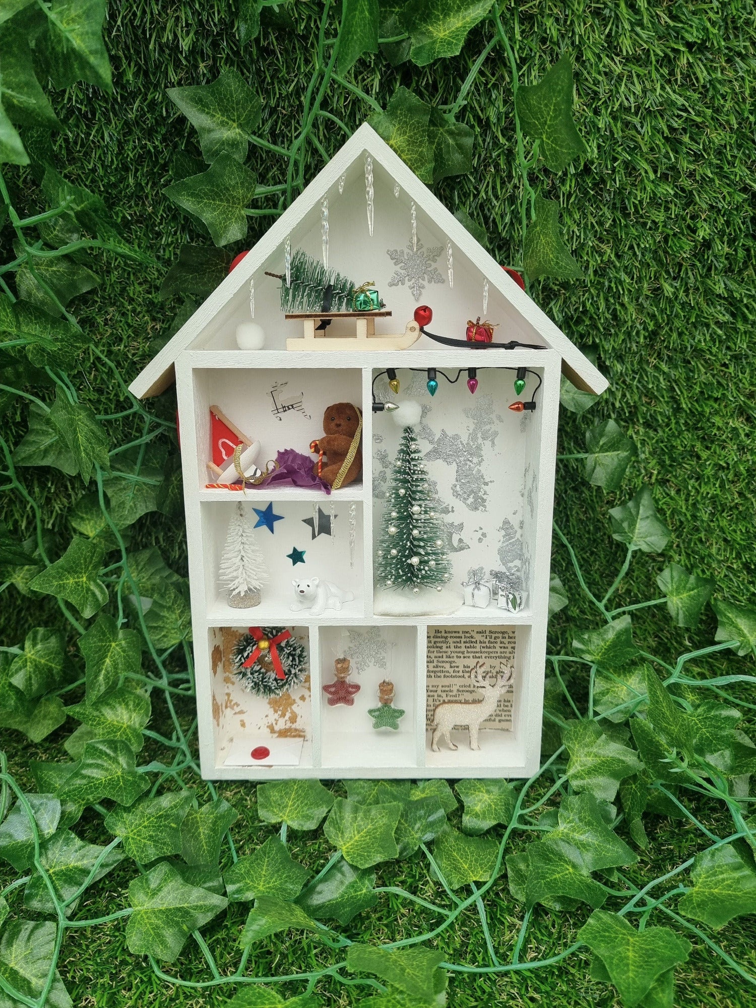 Small white shelving unit in the shape of a house with 7  compartments, each with a different Christmas scene including christmas tree, presents, baubles, wreath, stag, polar bear and sledge