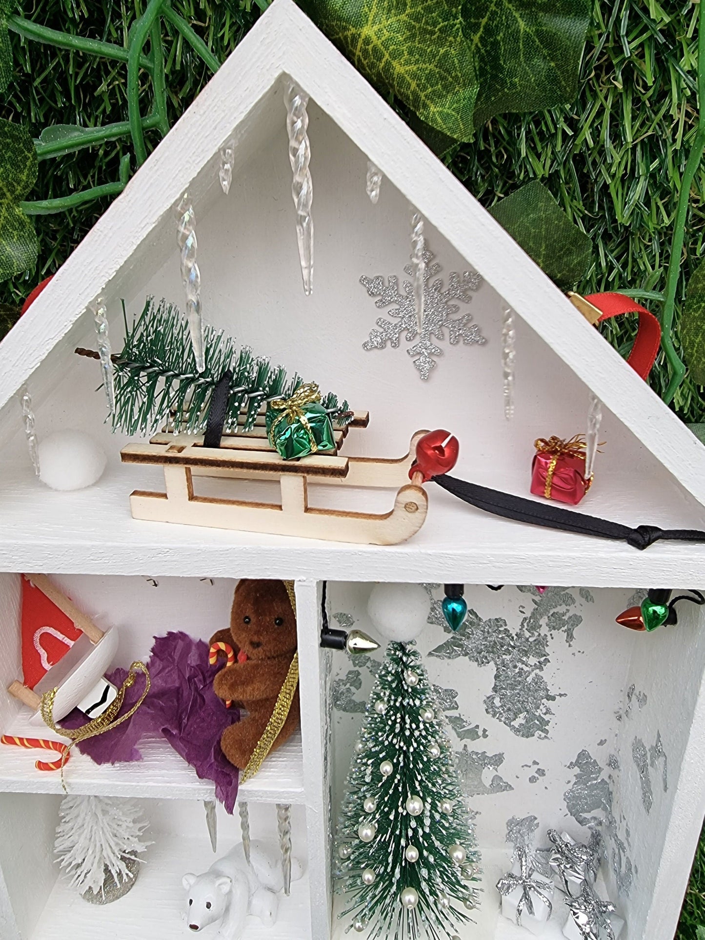 Detail of a small white shelving unit in the shape of a house. Each compartment has a different Christmas scene. There small sledge with a christmas tree. Icicles line the roof. A christmas tree and gifts are seen beneath