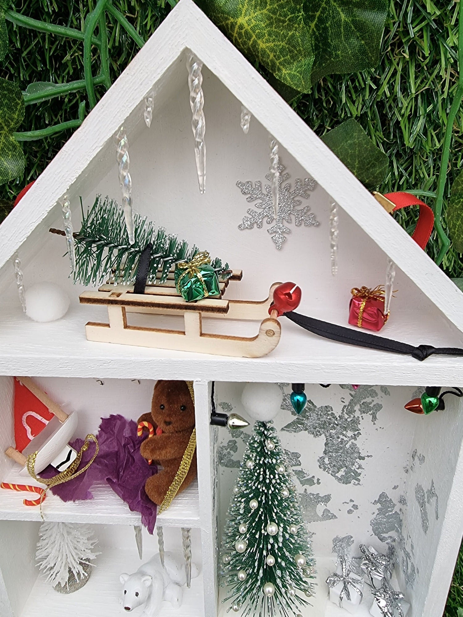 Detail of a small white shelving unit in the shape of a house. Each compartment has a different Christmas scene. There small sledge with a christmas tree. Icicles line the roof. A christmas tree and gifts are seen beneath