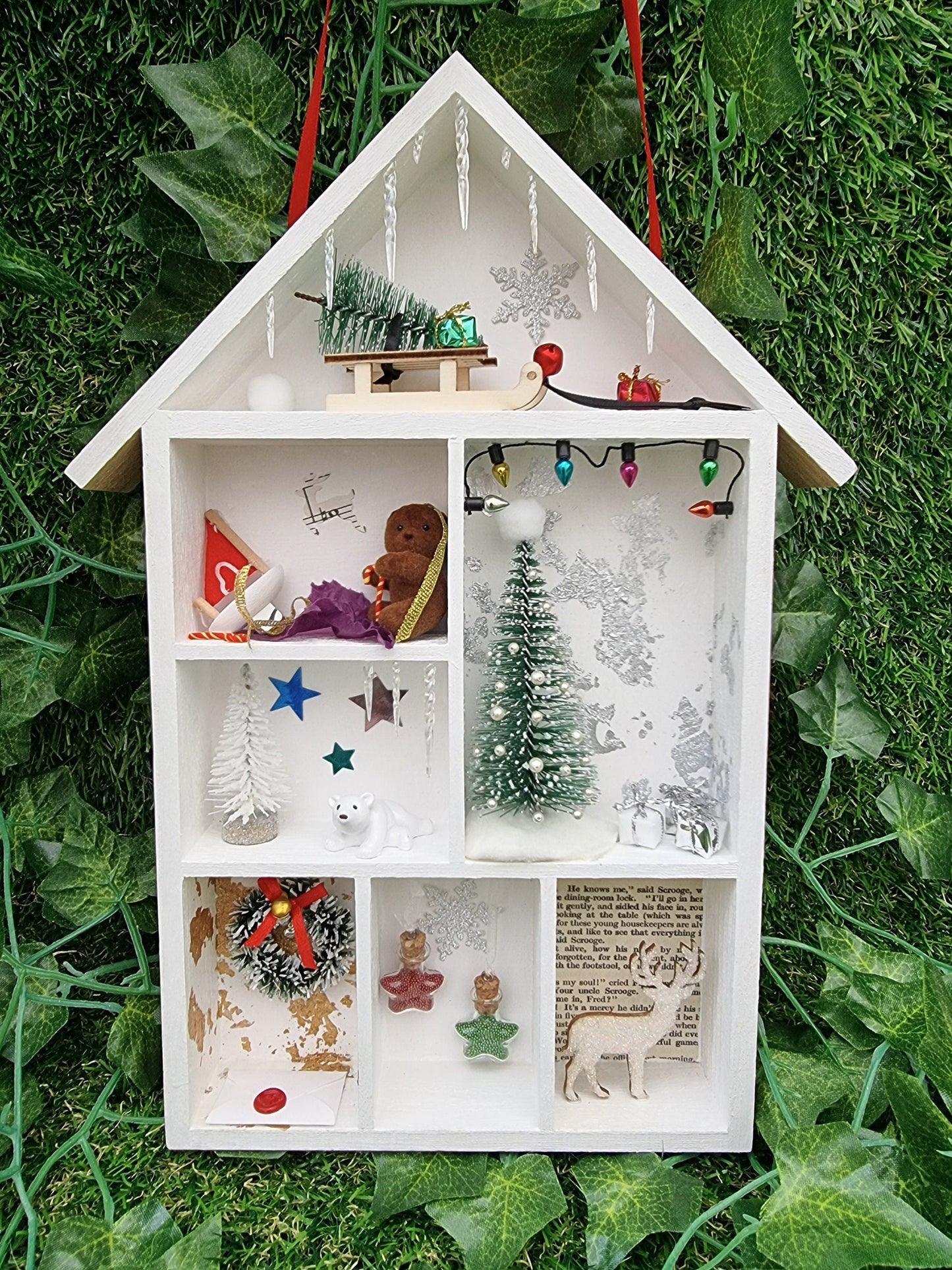 Small white shelving unit in the shape of a house with 7  compartments, each with a different Christmas scene including christmas tree, presents, baubles, wreath, stag, polar bear and sledge