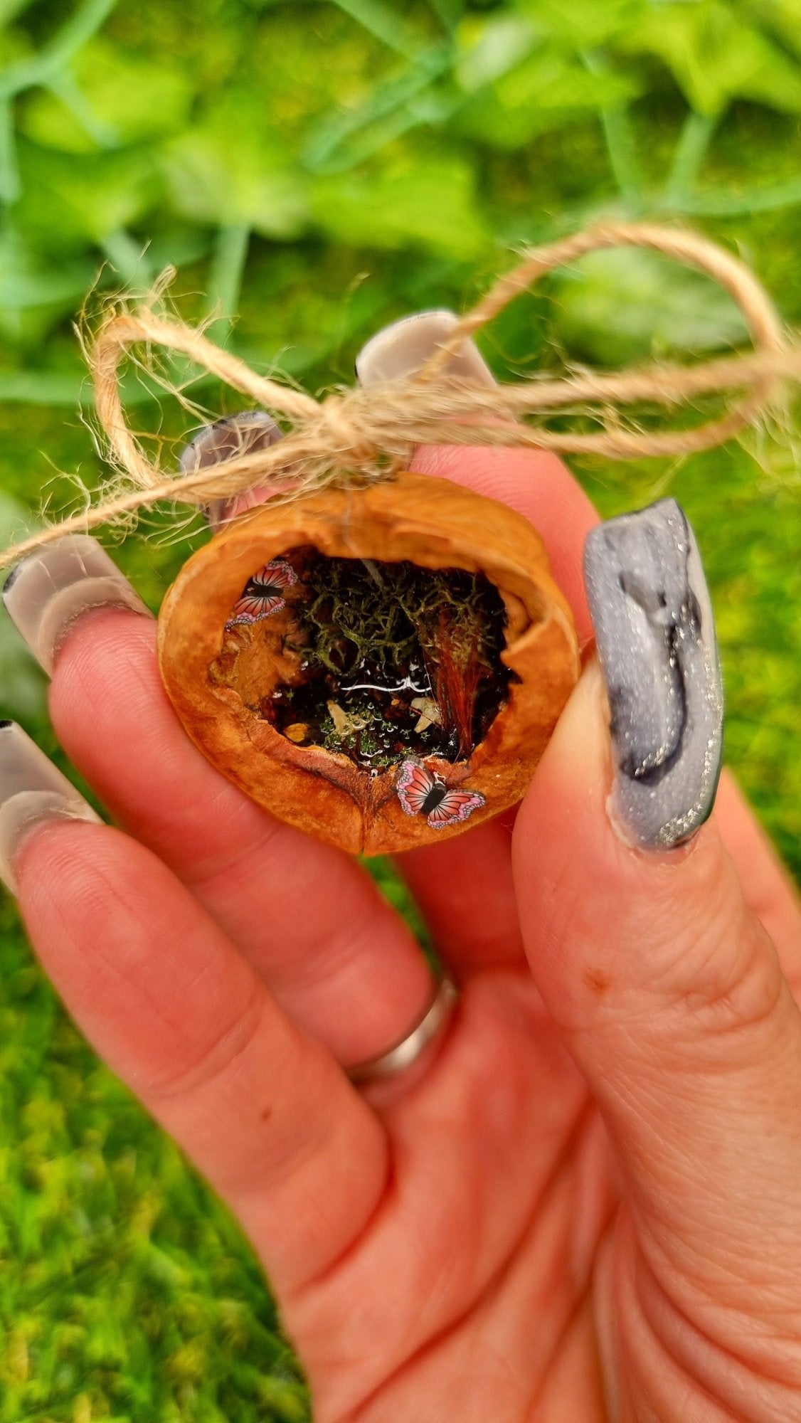 A hand holds a small brown walnut half shell containing a miniature diorama. The inside of the shell is natural brown with greenery, brown reeds and a tiny pond with leaves floating on the surface. There are two pink and purple butteflies