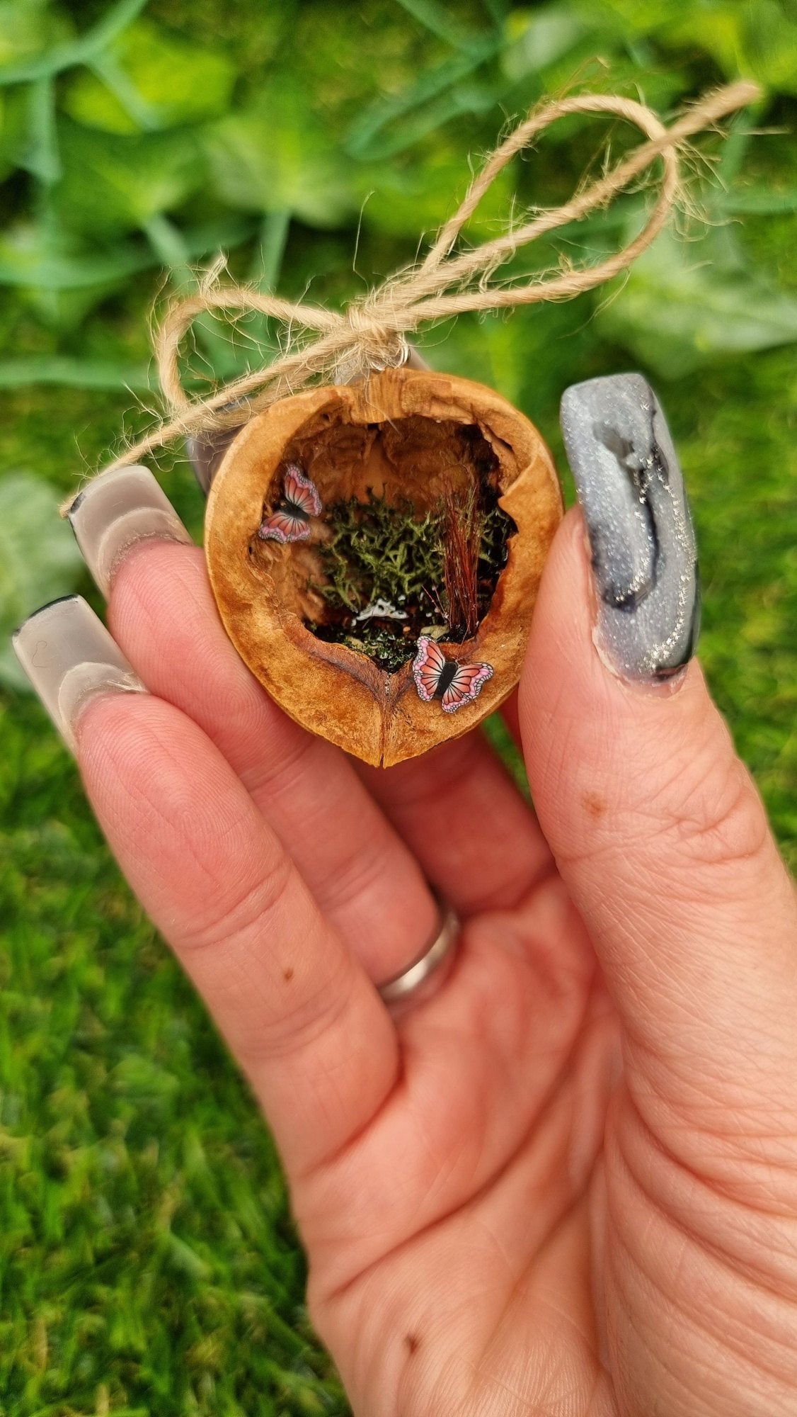 A hand holds a small brown walnut half shell containing a miniature diorama. The inside of the shell is natural brown with greenery, brown reeds and a tiny pond with leaves floating on the surface. There are two pink and purple butteflies