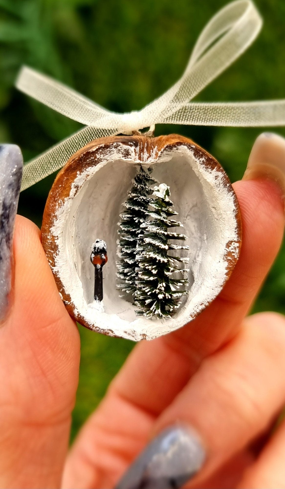 A hand holds a small walnut half shell containing a miniature diorama. The inside of the shell is snowy and white with 2 tiny pine trees edged in snow and a tiny black street lamp with an orange glow. The shell top has a cream organza ribbon in a bow