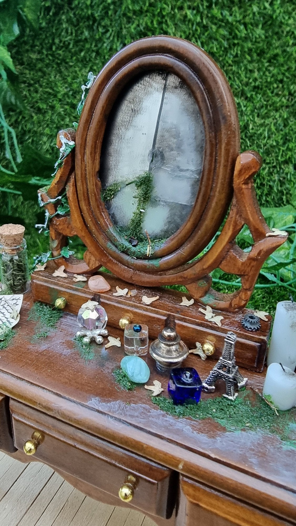 A 1:12 brown abandoned dressing table sits on a wooden base. It has a a broken mirror with moss in the breaks. The table top is strewn vessels, candles, leaves and moss. Ivy can be seen climbing up the left support of the mirror.