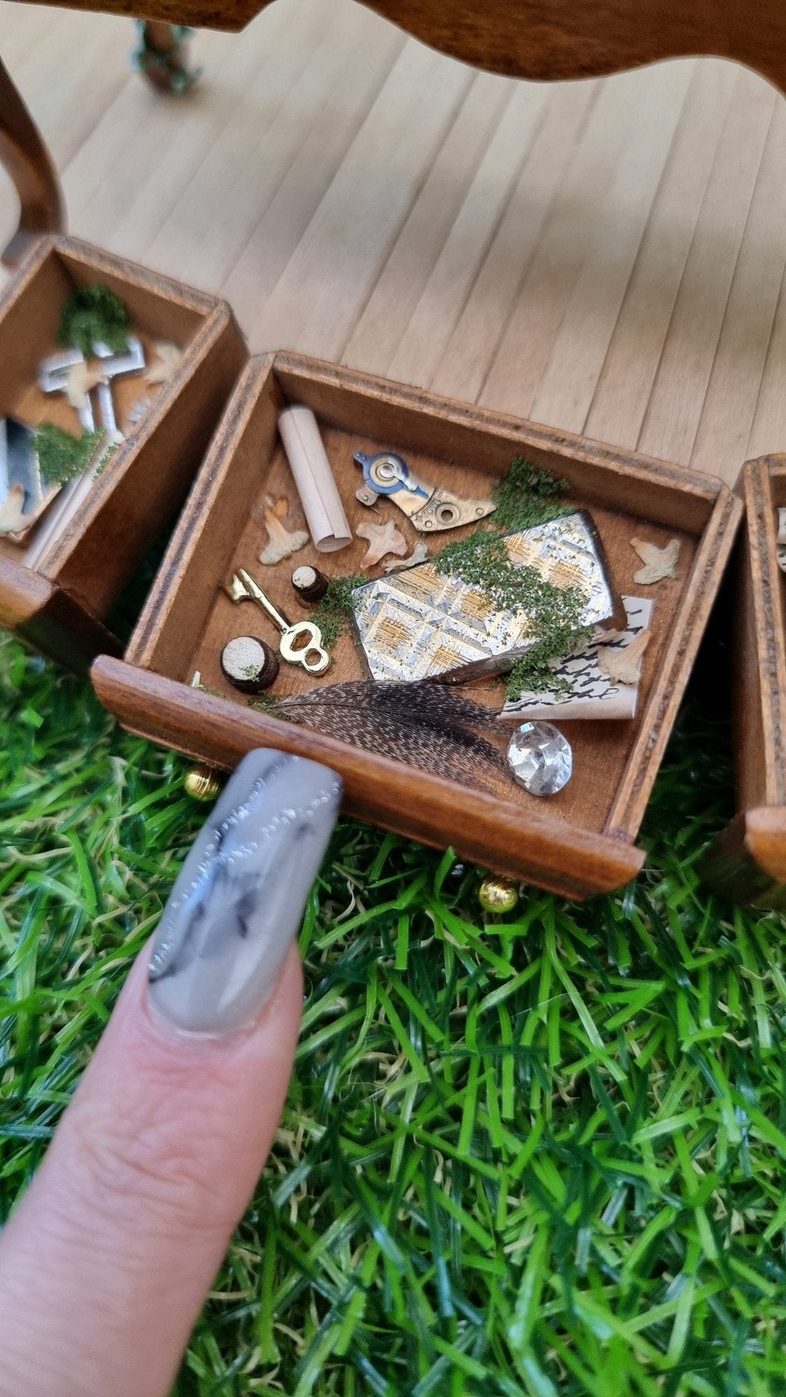 A finger points to an open drawer of a 1:12 abandoned dressing table. Inside is a feather, a patterned metal box, jewel, written note, key, navigation tool, moss and leaves.