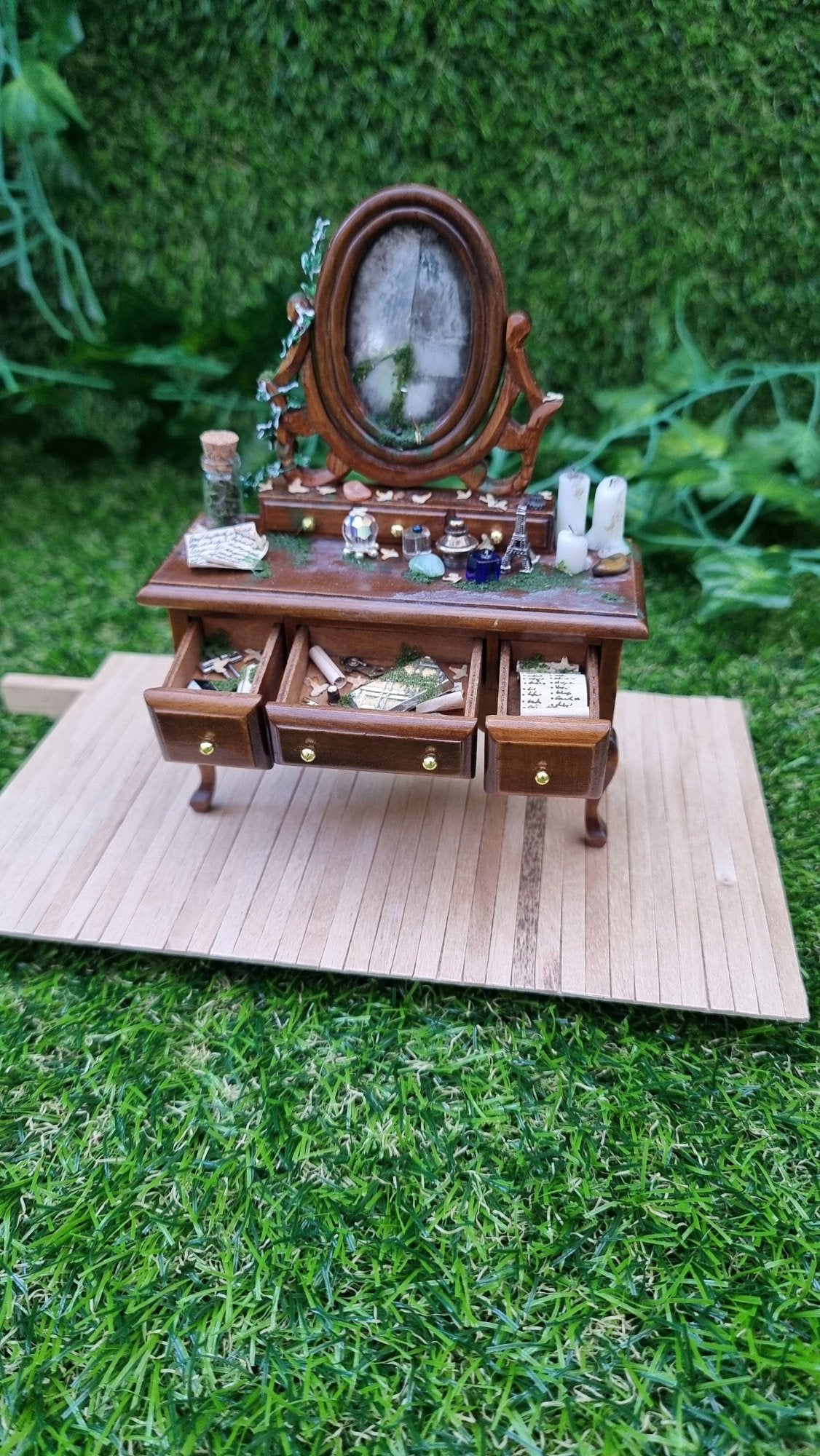 A 1:12 brown abandoned dressing table sits on a wooden base with 3 drawers open. The dressing table has a broken mirror and the table top is strewn vessels, candles and moss. The open drawers are filled with curios and papers