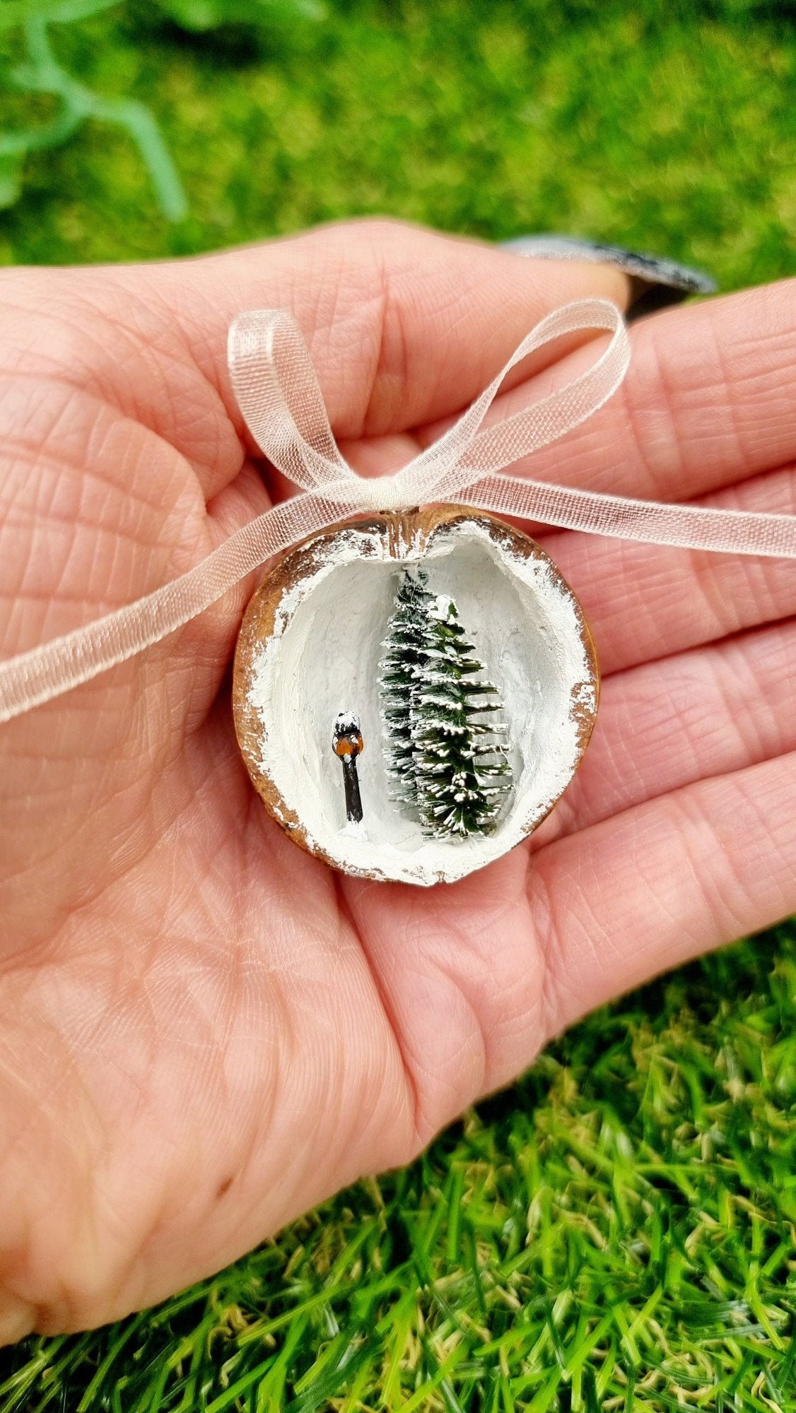 A hand holds a small walnut half shell containing a miniature diorama. The inside of the shell is snowy and white with 2 tiny pine trees edged in snow and a tiny black street lamp with an orange glow. The shell top has a cream organza ribbon in a bow