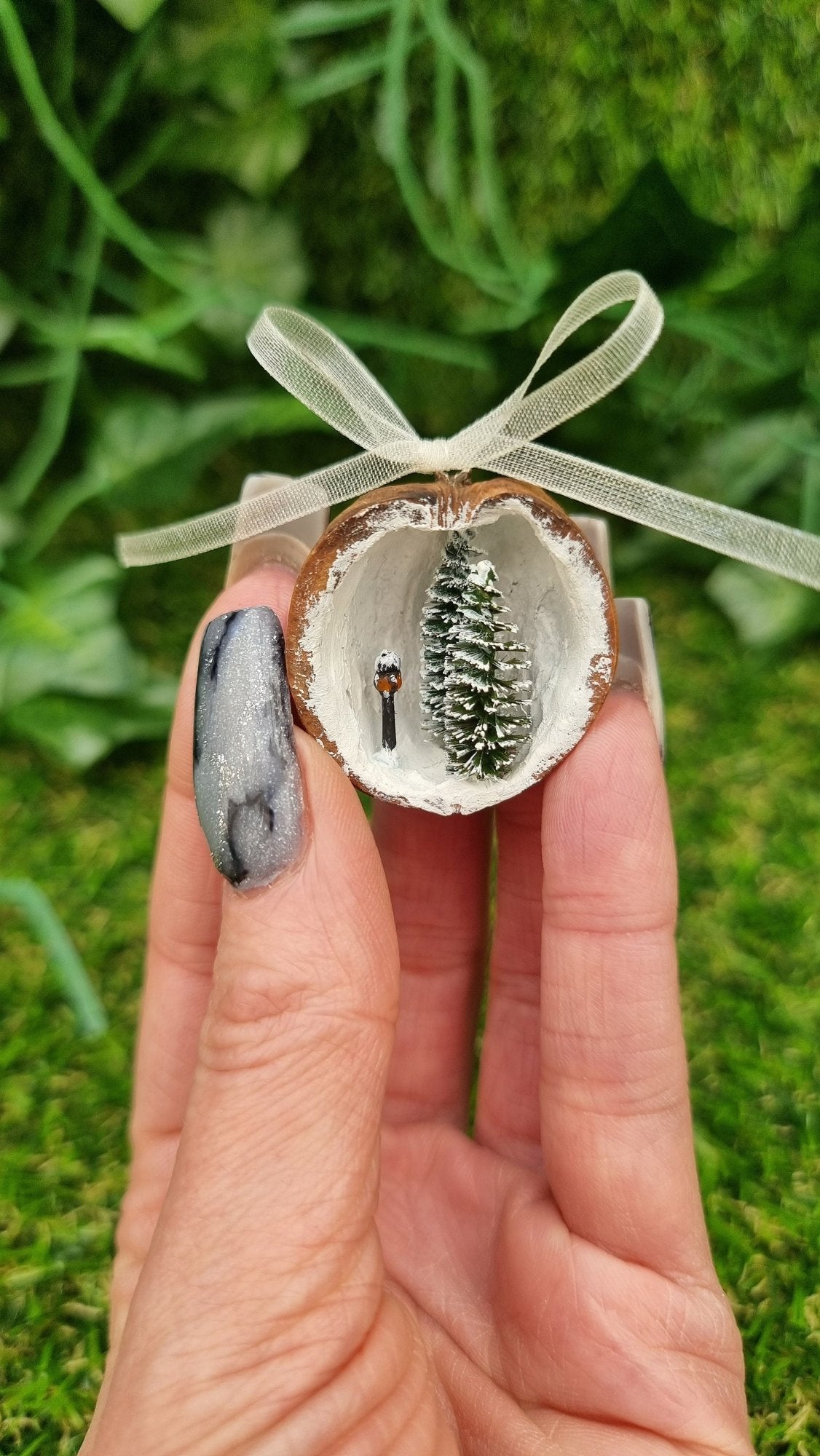 A hand holds a small walnut half shell containing a miniature diorama. The inside of the shell is snowy and white with 2 tiny pine trees edged in snow and a tiny black street lamp with an orange glow. The shell top has a cream organza ribbon in a bow