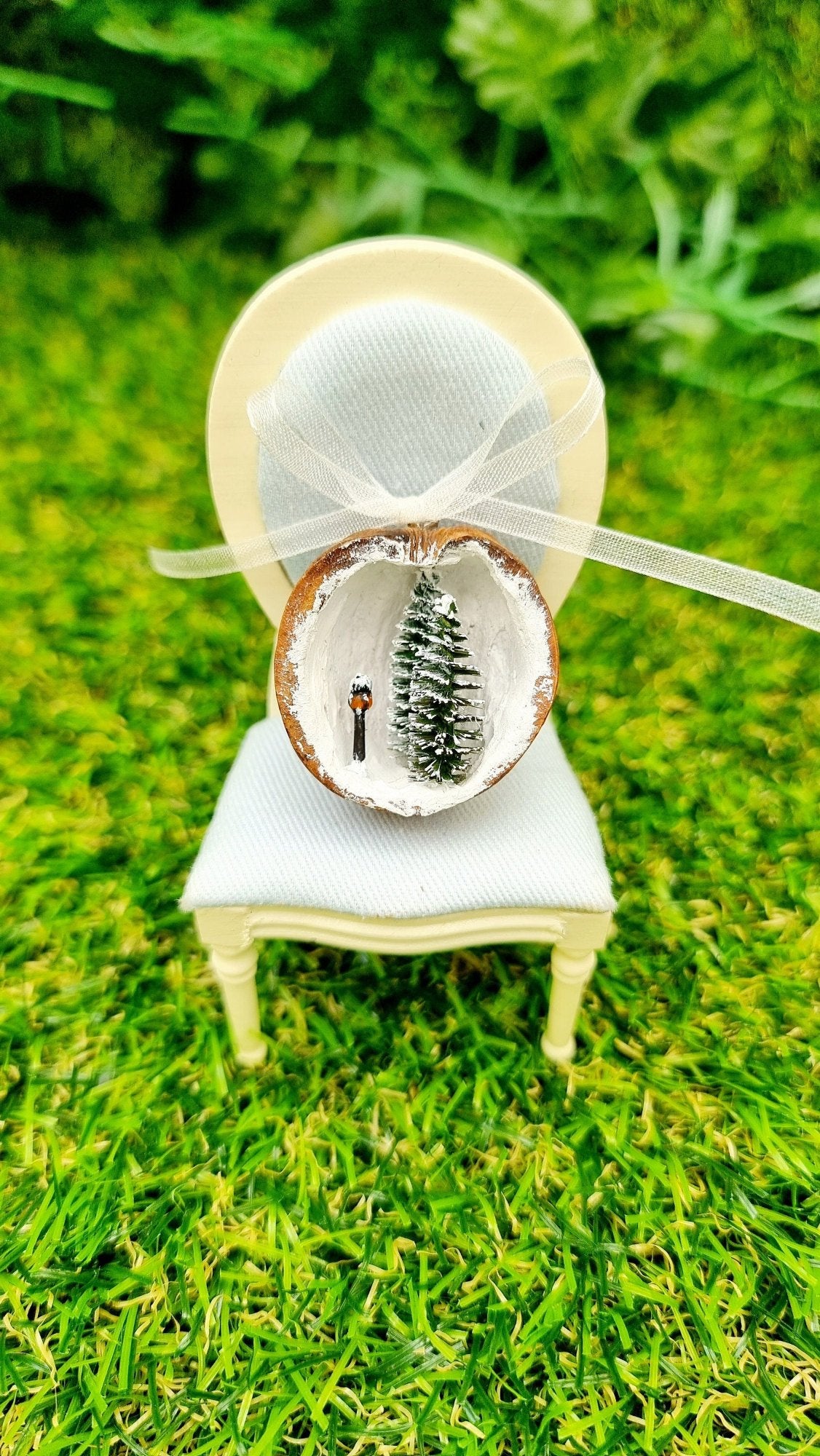 A walnut half shell containing a miniature diorama sits on a 1:12 scale chair. The inside of the shell is snowy and white with 2 tiny pine trees edged in snow and a tiny black street lamp with an orange glow. The shell is topped with a bow