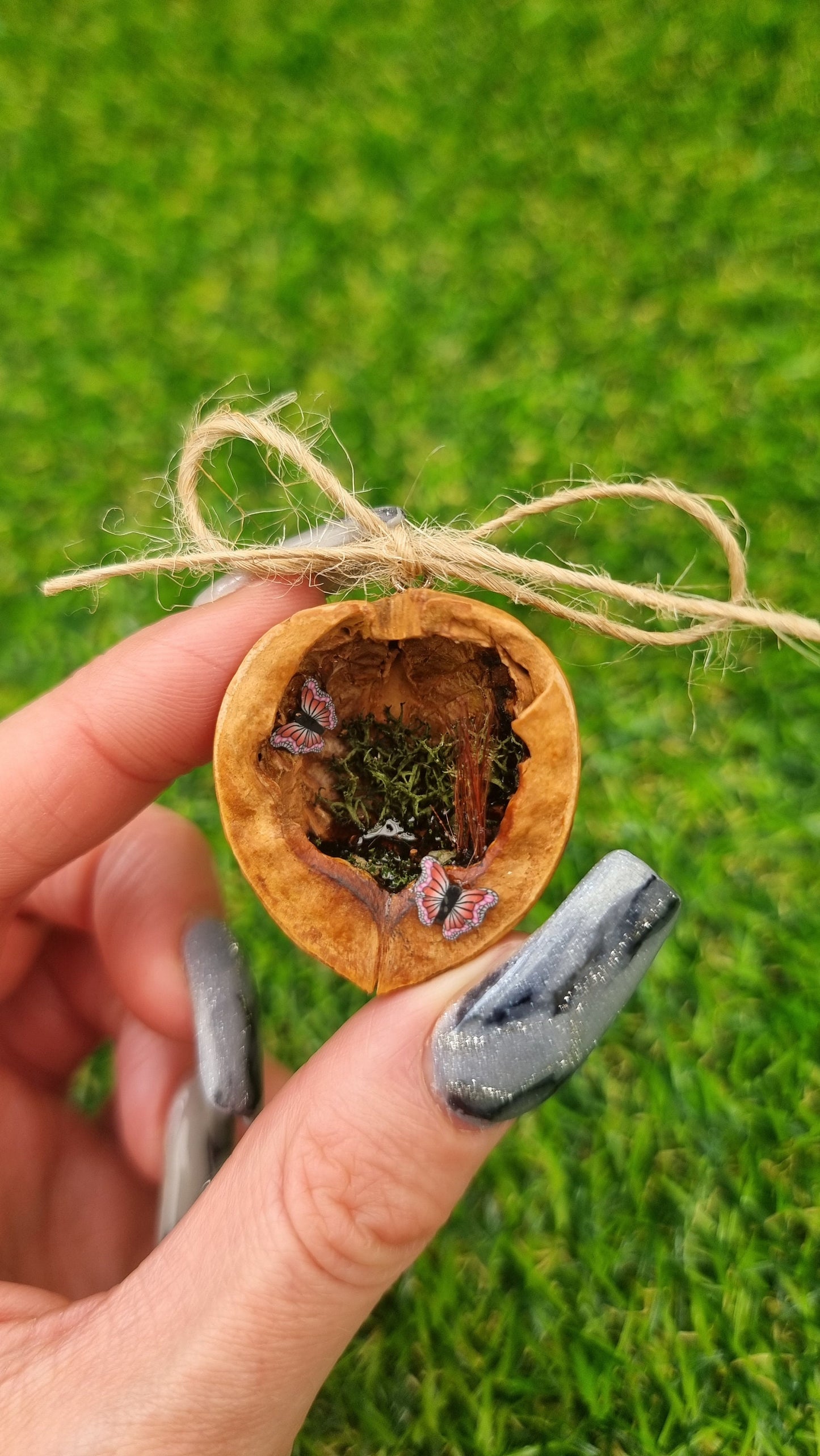 A hand holds a small brown walnut half shell containing a miniature diorama. The inside of the shell is natural brown with greenery, brown reeds and a tiny pond with leaves floating on the surface. There are two pink and purple butteflies