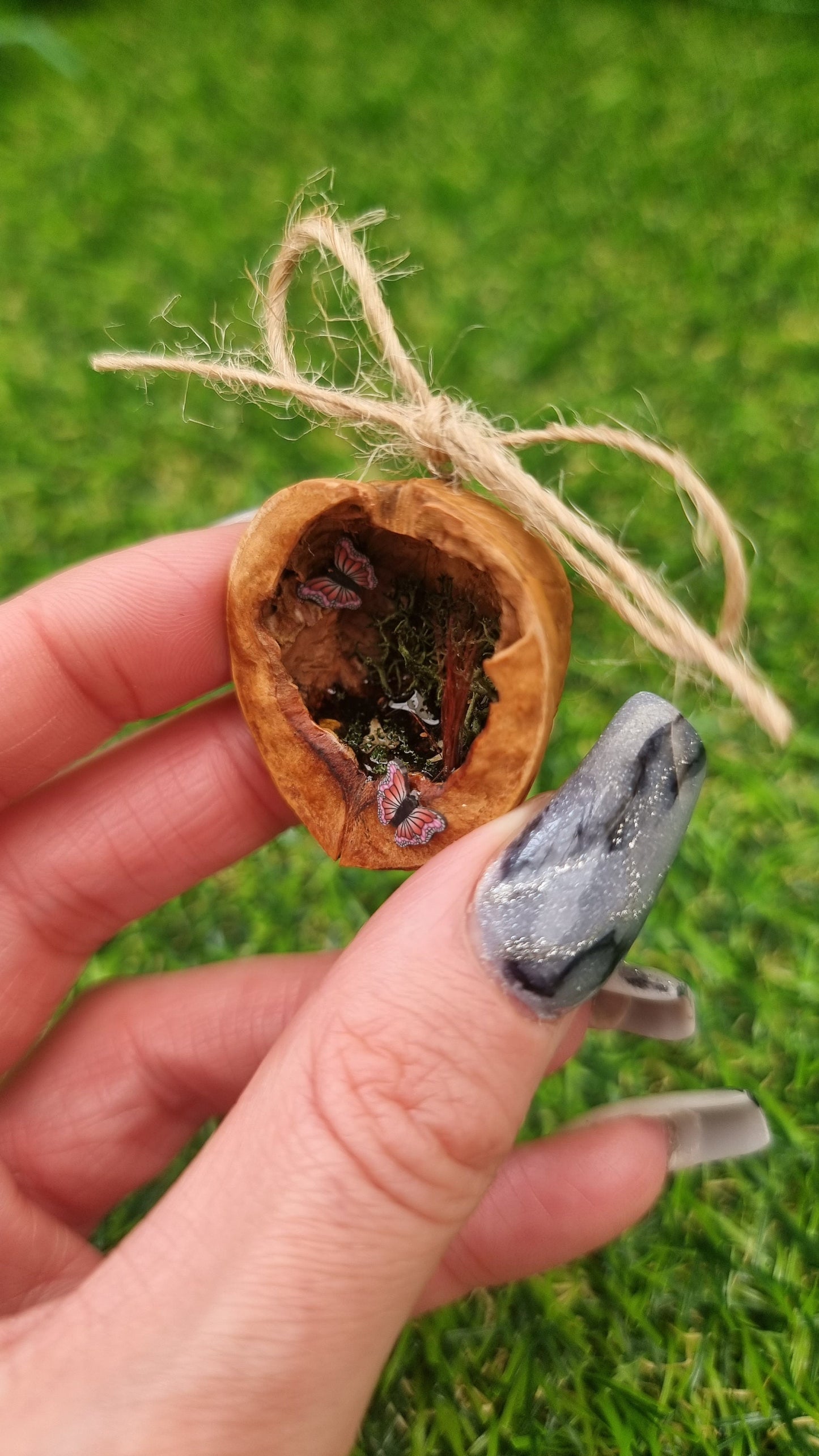 A hand holds a small brown walnut half shell containing a miniature diorama. The inside of the shell is natural brown with greenery, brown reeds and a tiny pond with leaves floating on the surface. There are two pink and purple butteflies