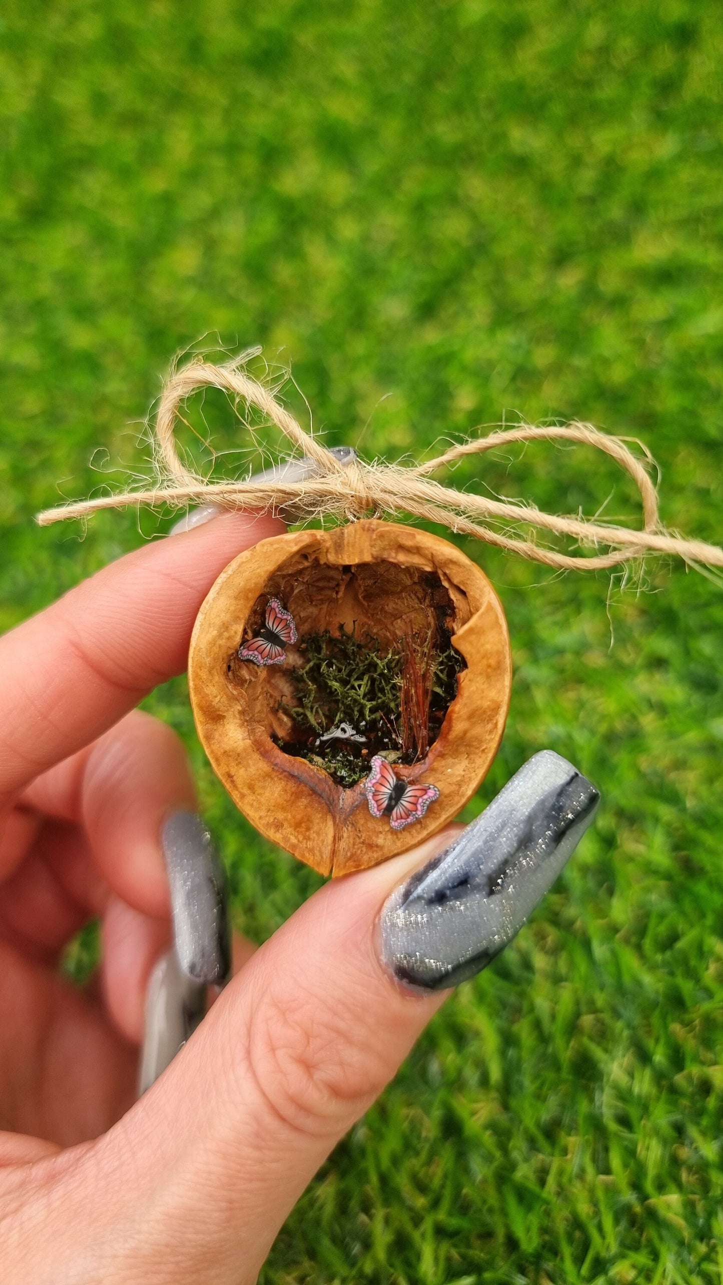 A hand holds a small brown walnut half shell containing a miniature diorama. The inside of the shell is natural brown with greenery, water and two pink and purple butteflies- one inside and one on the edge. The shell top has a jute string bow