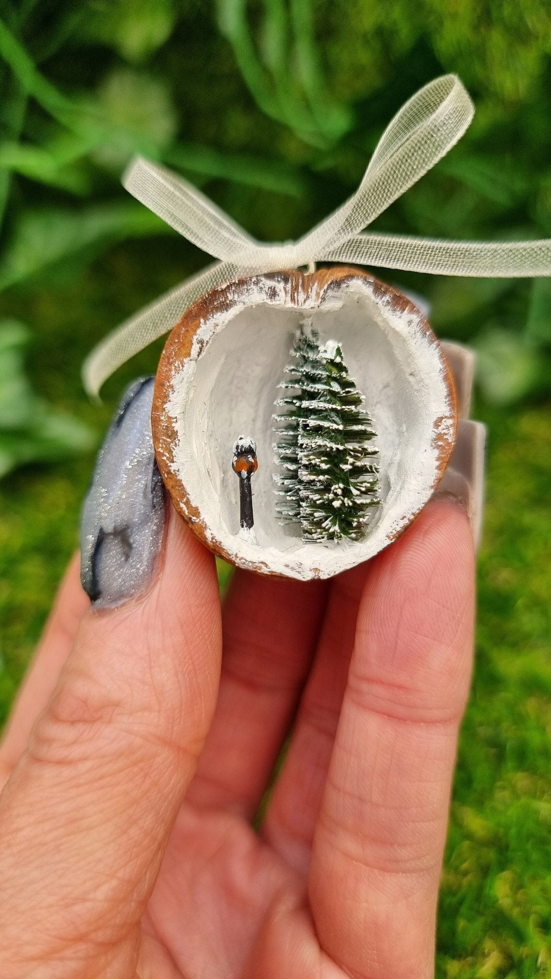 A hand holds a small walnut half shell containing a miniature diorama. The inside of the shell is snowy and white with 2 tiny pine trees edged in snow and a tiny black street lamp with an orange glow. The shell top has a cream organza ribbon in a bow