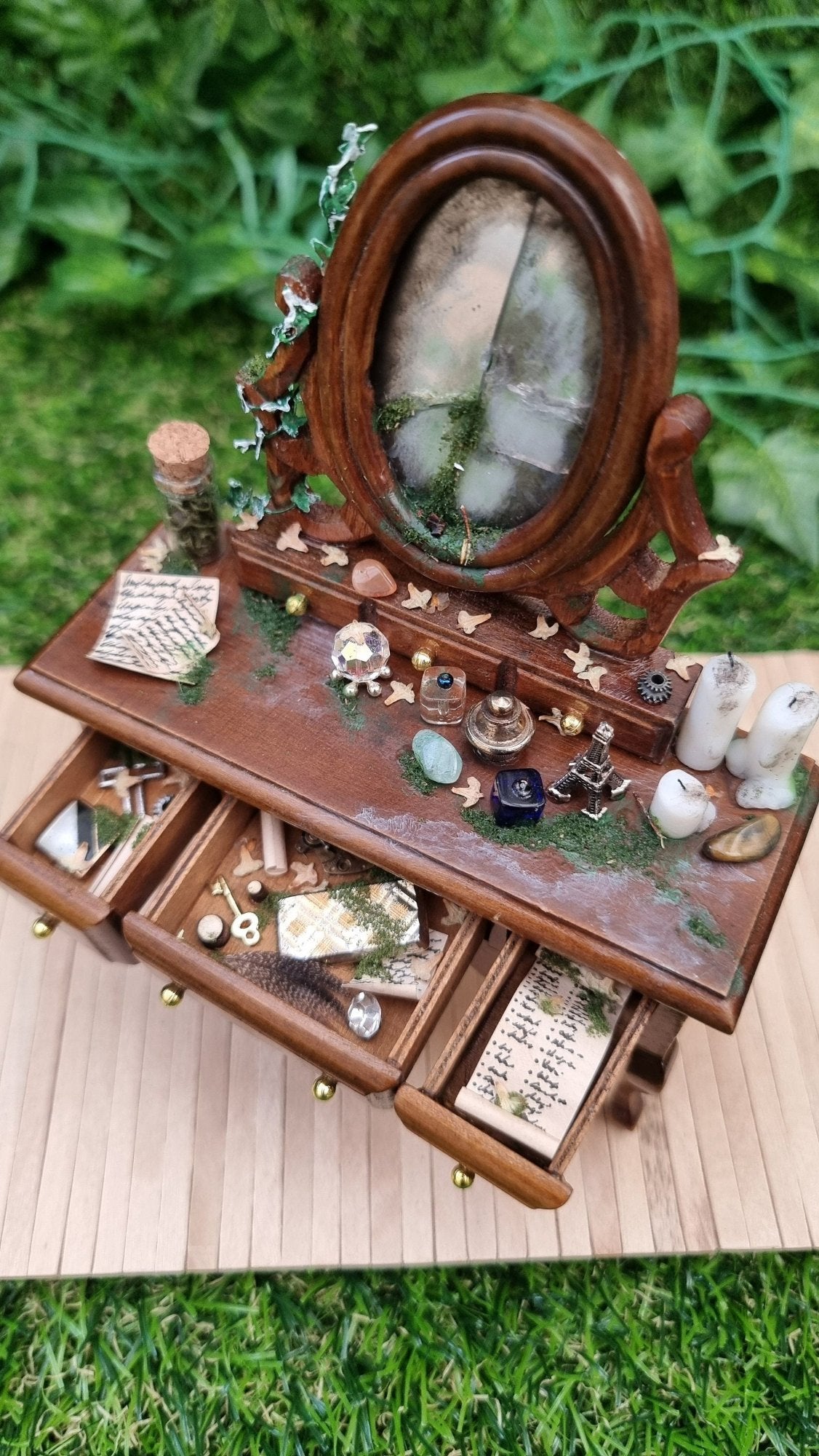 A 1:12 brown abandoned dressing table sits on a wooden base with 3 drawers open. The dressing table has a broken mirror and the table top is strewn vessels, candles and moss. The open drawers are filled with curios and papers