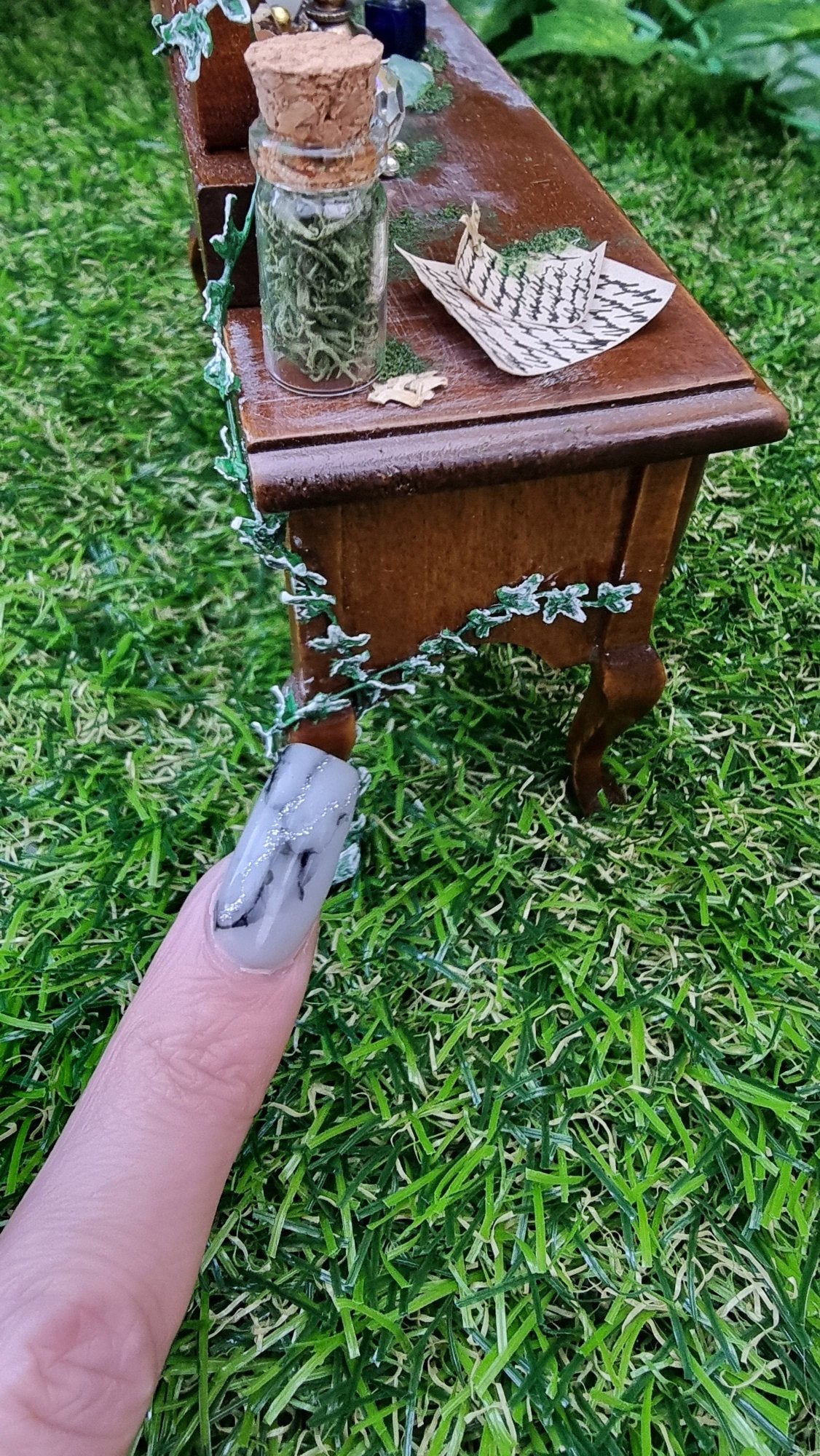 A finger points to miniature ivy climbing up the back of a 1:12 abandoned dressing table, a glass bottle and handwritten notes sit on the dressing table top