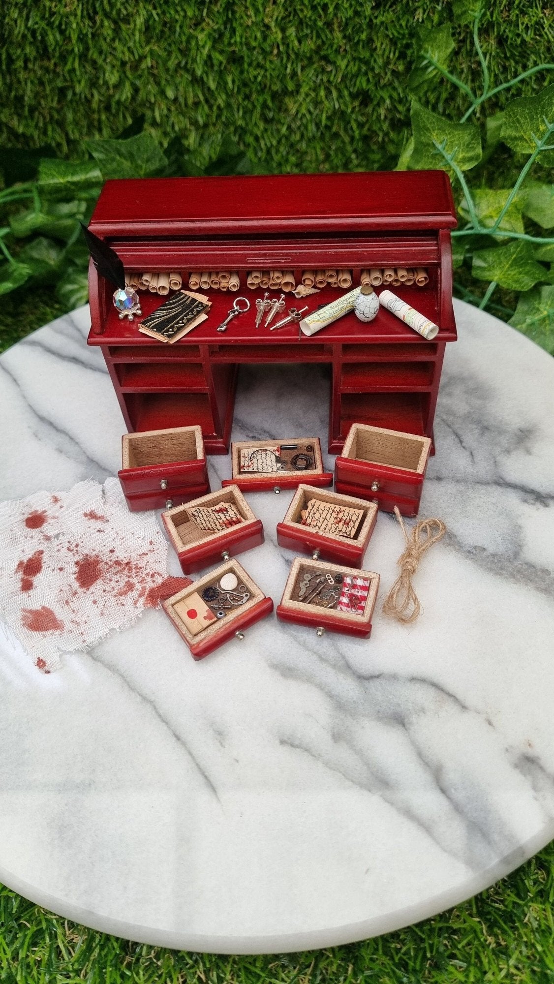 1:12 scale rolltop desk sits on a marble slab. The desk is open and has 7 filled drawers laid out in front of it. There is also a loop of rope and a bloody rag. The desk top has maps, key, syringes, journal inkwell and quill