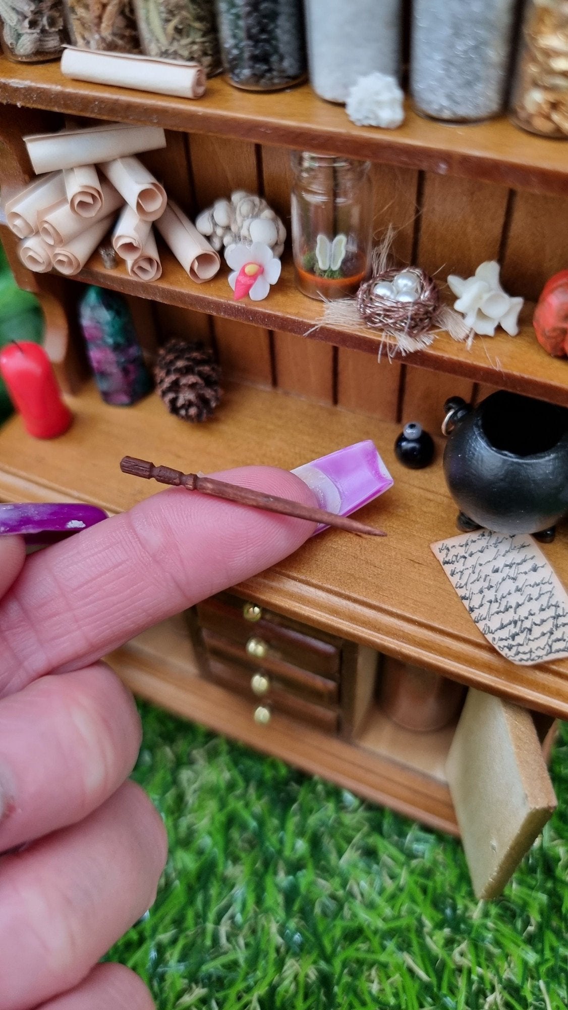 A 1:12 miniature brown dresser is shown with 2 shelves with filled bottles, parchment rolls and curios. The counter top has a candle and cauldron. A tiny want sits on a finger for scale.