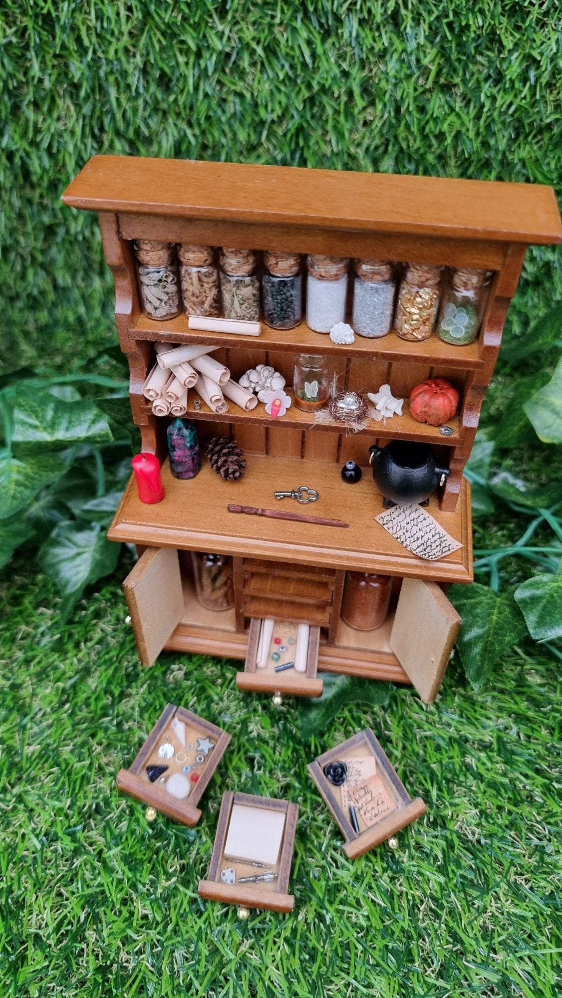 A 1:12 miniature brown dresser has 2 shelves with filled bottles, parchment rolls and curios. The counter top has a candle and cauldron. There are 2 open cupboard doors with bottles and 4  drawers laid in front notes and curios.