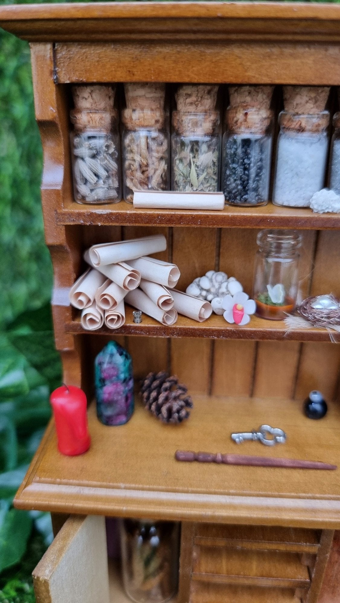 A 1:12 miniature brown dresser is shown with 2 shelves with filled bottles, parchment rolls and curios. The counter top has a ruby zoisite point, candle pine cone key and wand.