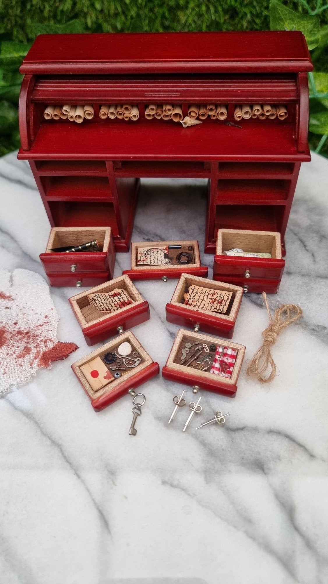 1:12 scale rolltop desk sits on a marble slab. The desk is open and has 7 filled drawers laid out in front of it. There is also a loop of rope, a bloody rag, maps, key, syringes, journal inkwell and quill