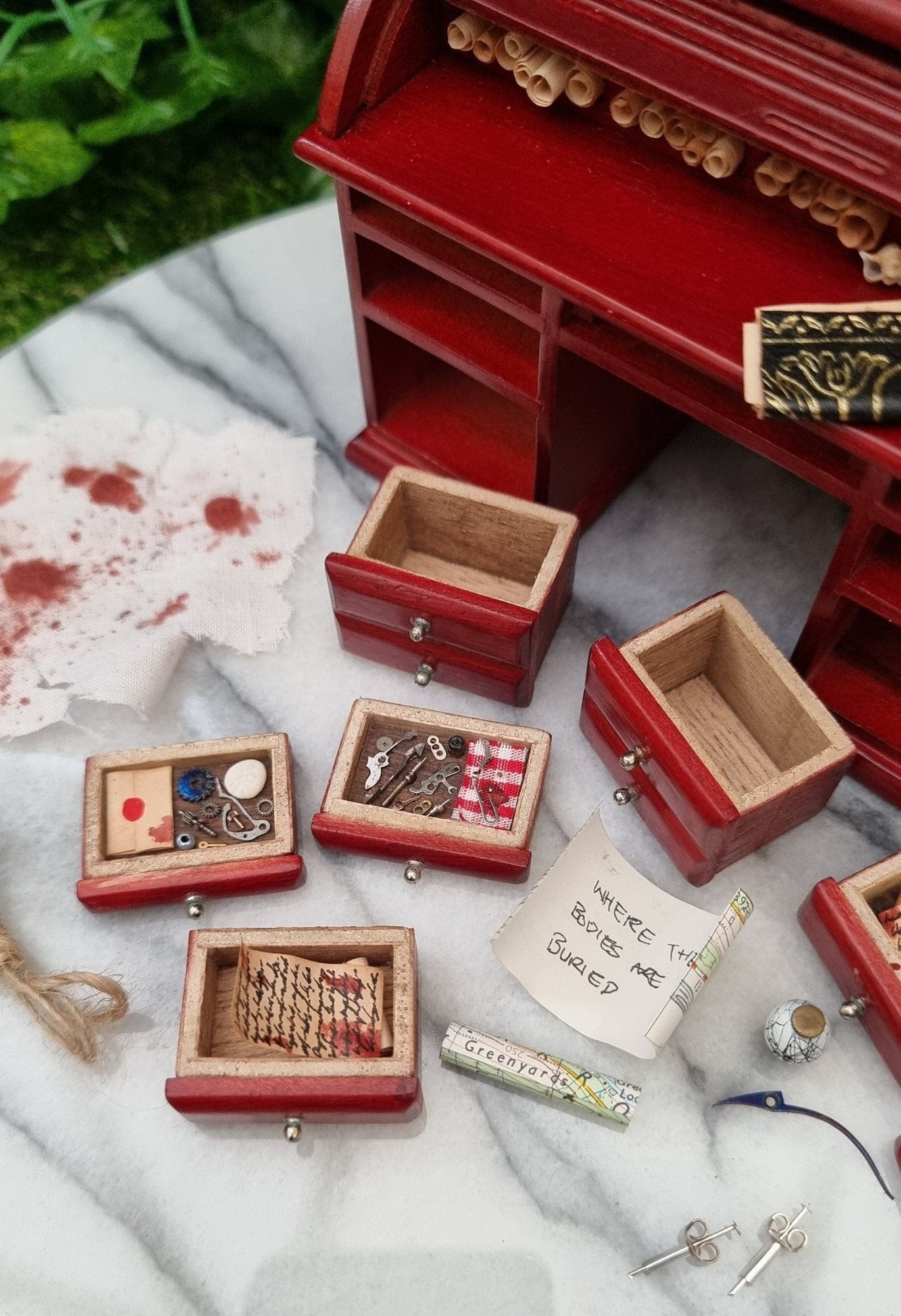 1:12 scale rolltop desk sits on a marble slab. The desk is open and has 6 drawers laid out in front of it. There is also a loop of rope, a bloody ra, key, syringes and a map scroll. The back of one map reads Where the bodiesare buried