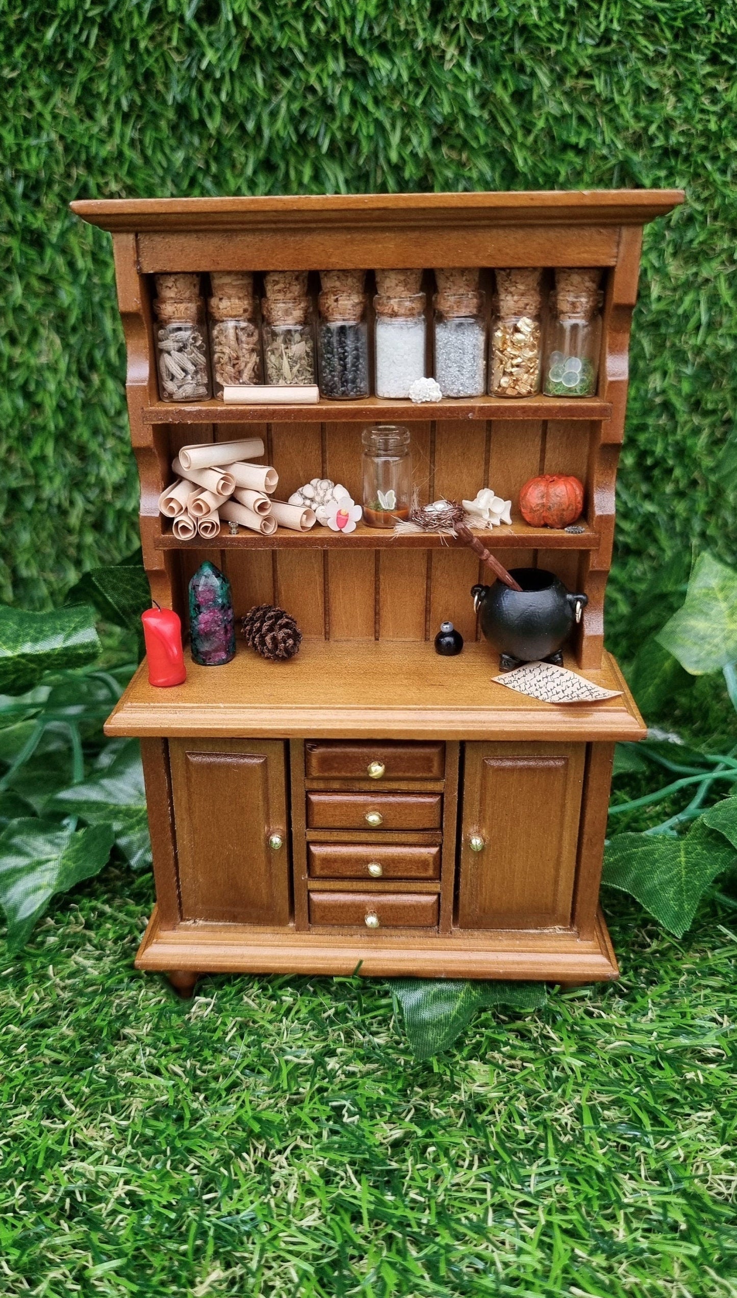 A miniature brown dresser is shown against a green background. The dresser has 2 shelves with filled bottles, parchment rolls and curios. The counter top has a candle and cauldron. There are 2 cupboard doors and 4 drawers